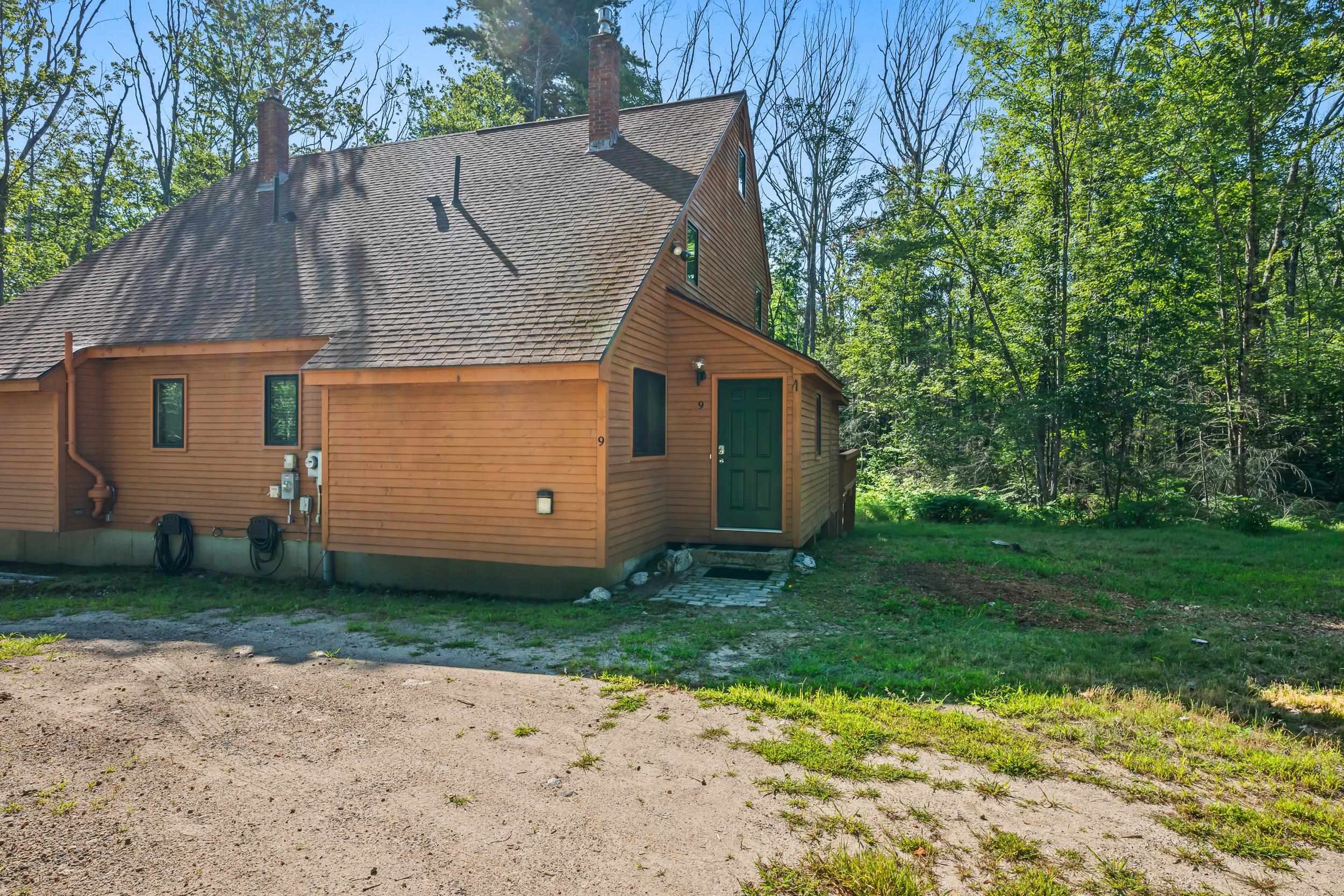 Brown wood-sided home with a green front door beside a wooded lawn and gravel driveway.