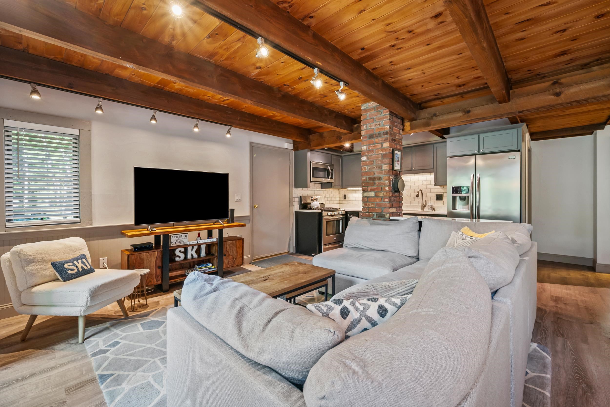 Open-plan living room and kitchen with a gray sectional, exposed wood beams, and a brick column.