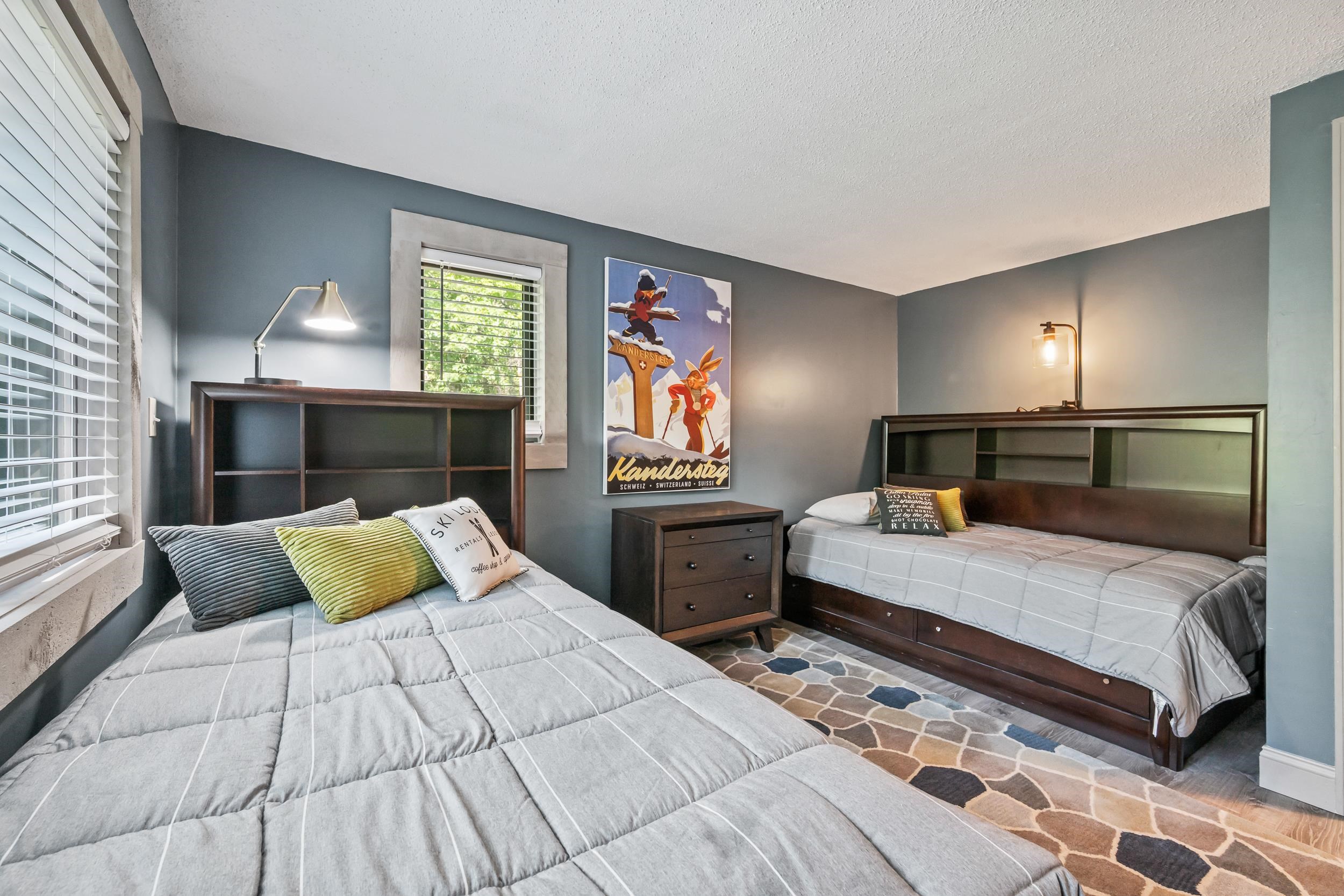 Bedroom with two single beds, dark wood headboards, gray bedding, and a colorful Kandersteg poster.