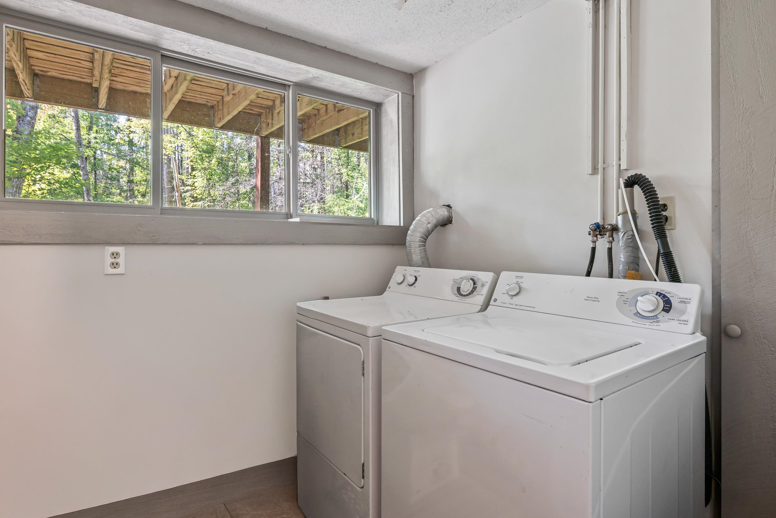 Laundry room with a washer and dryer beside a wide window overlooking trees.