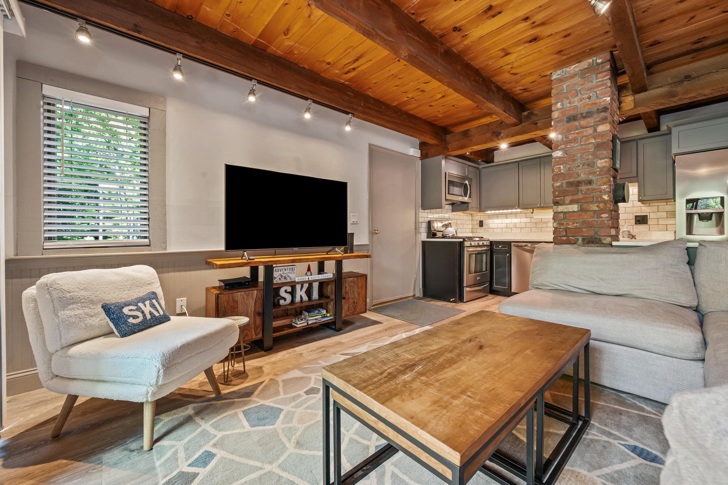 Furnished living room and kitchen with vaulted wood ceiling, brick column, and gray sectional.
