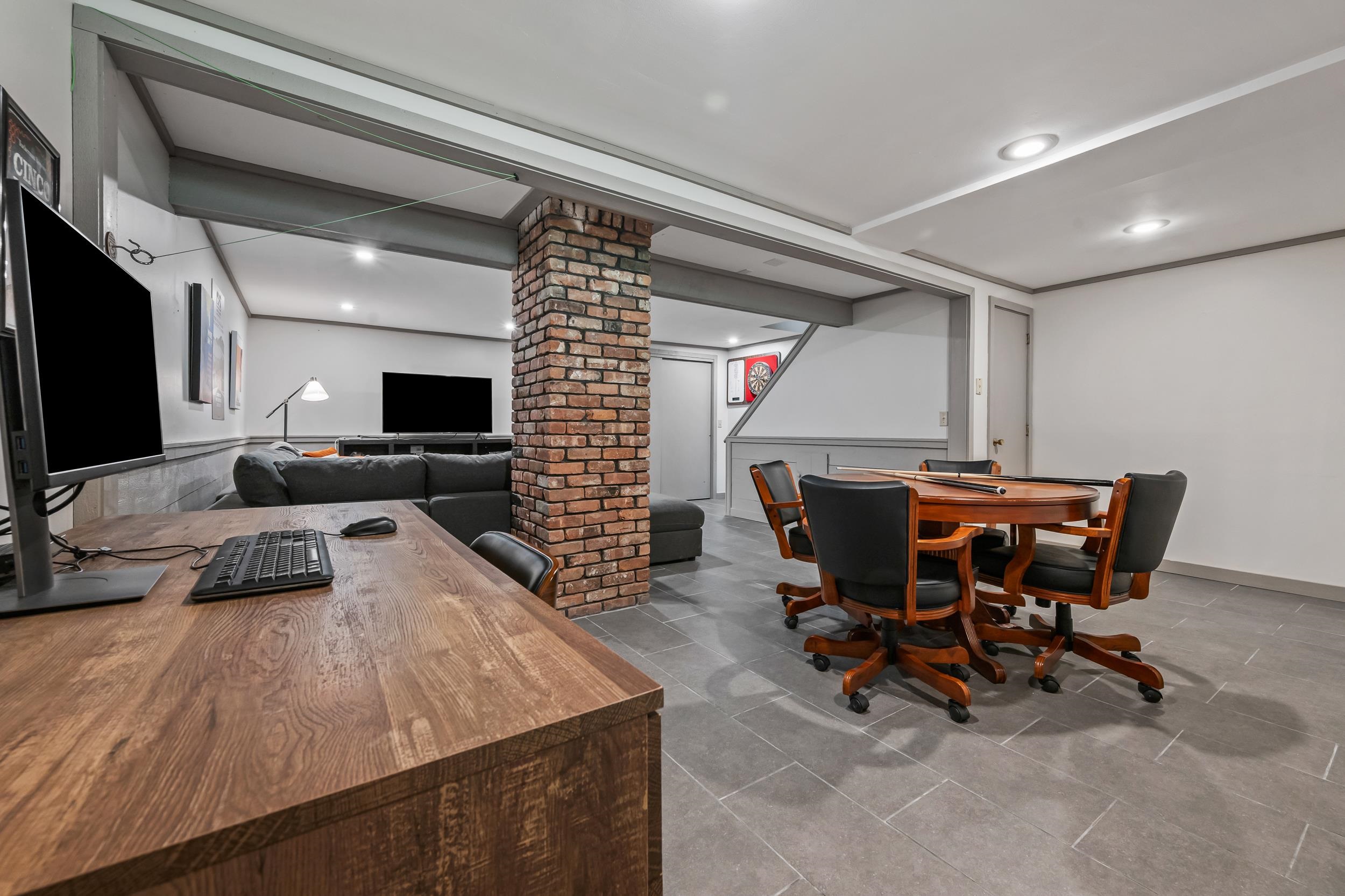 Basement recreation room with a brick pillar, sectional sofa, desk, televisions, and a game table.