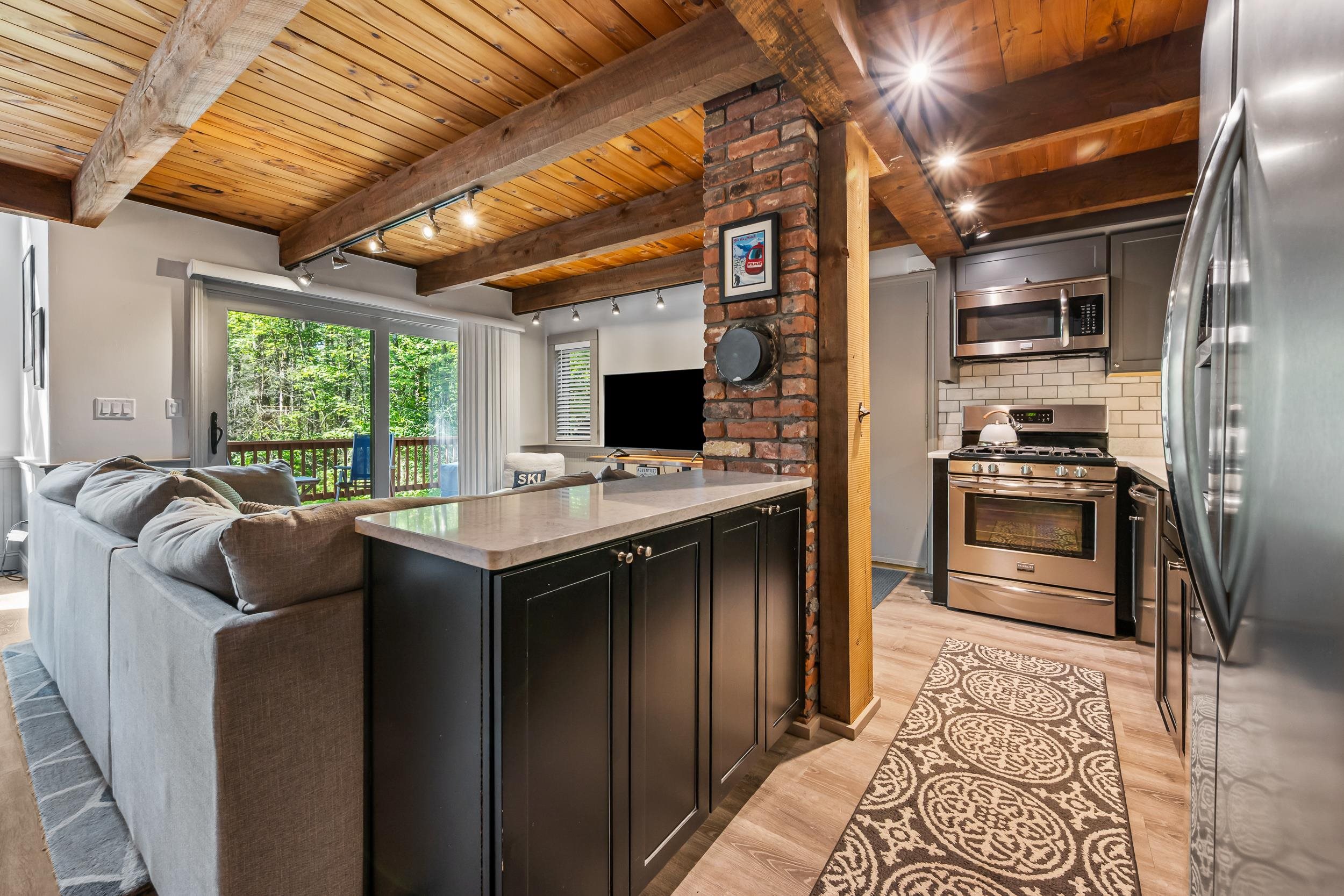 Open kitchen and living area with a vaulted wood ceiling, brick column, and sliding doors to a leafy deck.