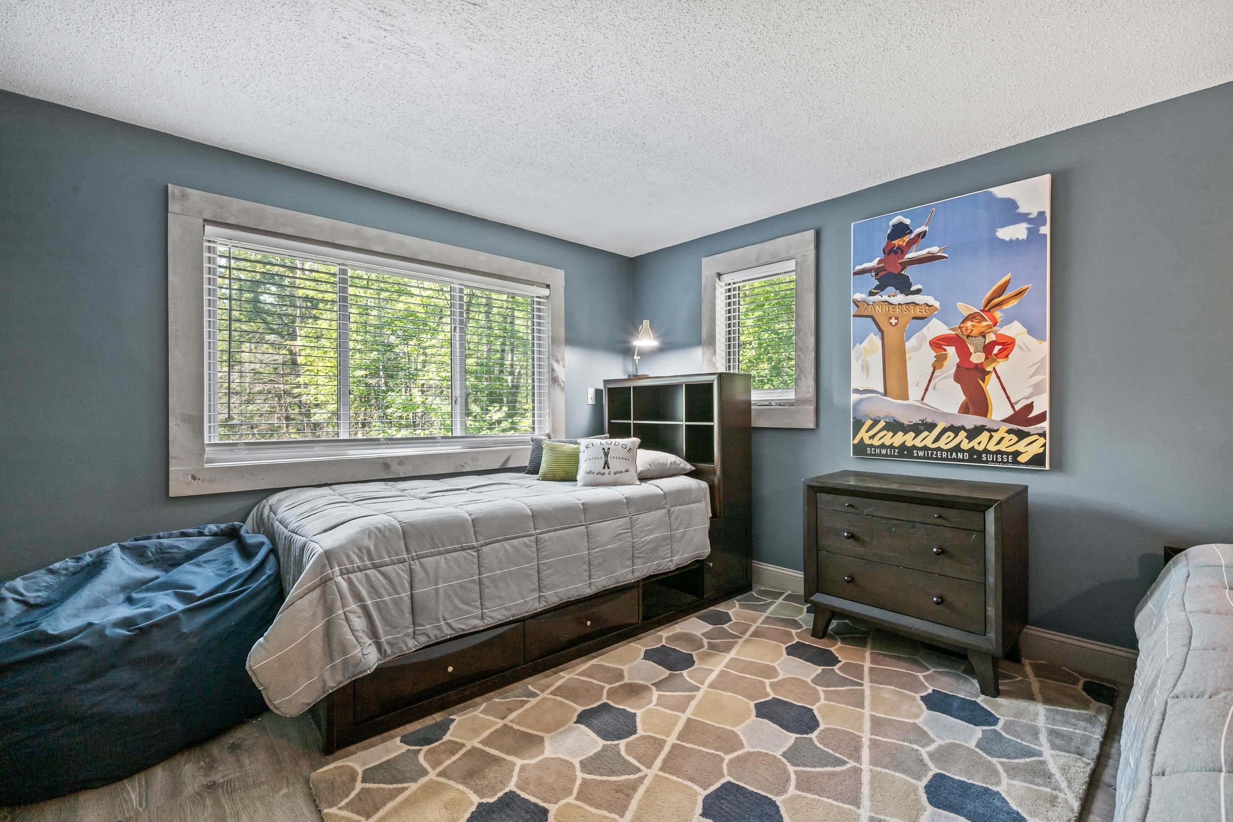 Furnished bedroom with a gray bed, leafy views through wide windows, and a vintage Kandersteg ski poster.