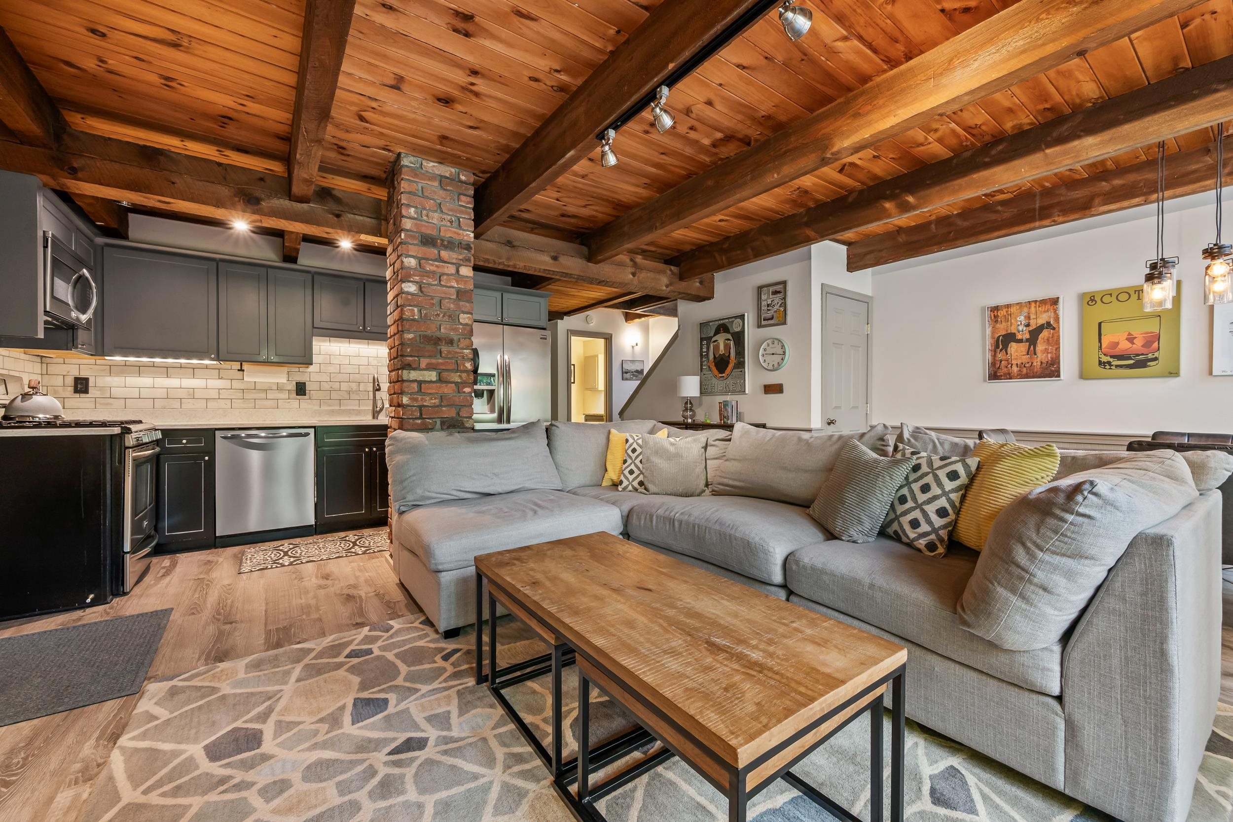 Open-plan living room and kitchen with a gray sectional, brick column, and exposed wood ceiling beams.
