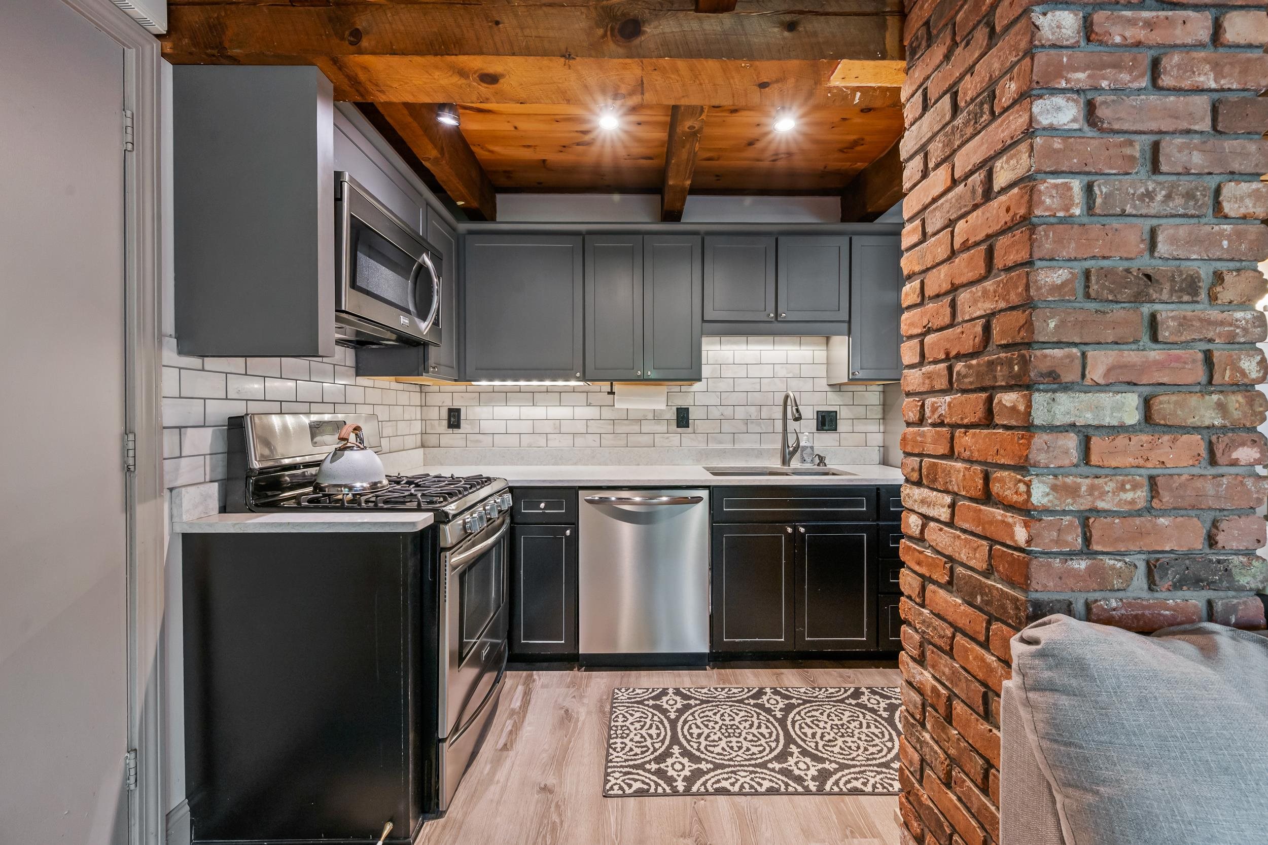 Kitchen with dark cabinets, stainless steel appliances, exposed wood ceiling beams, and a brick column.