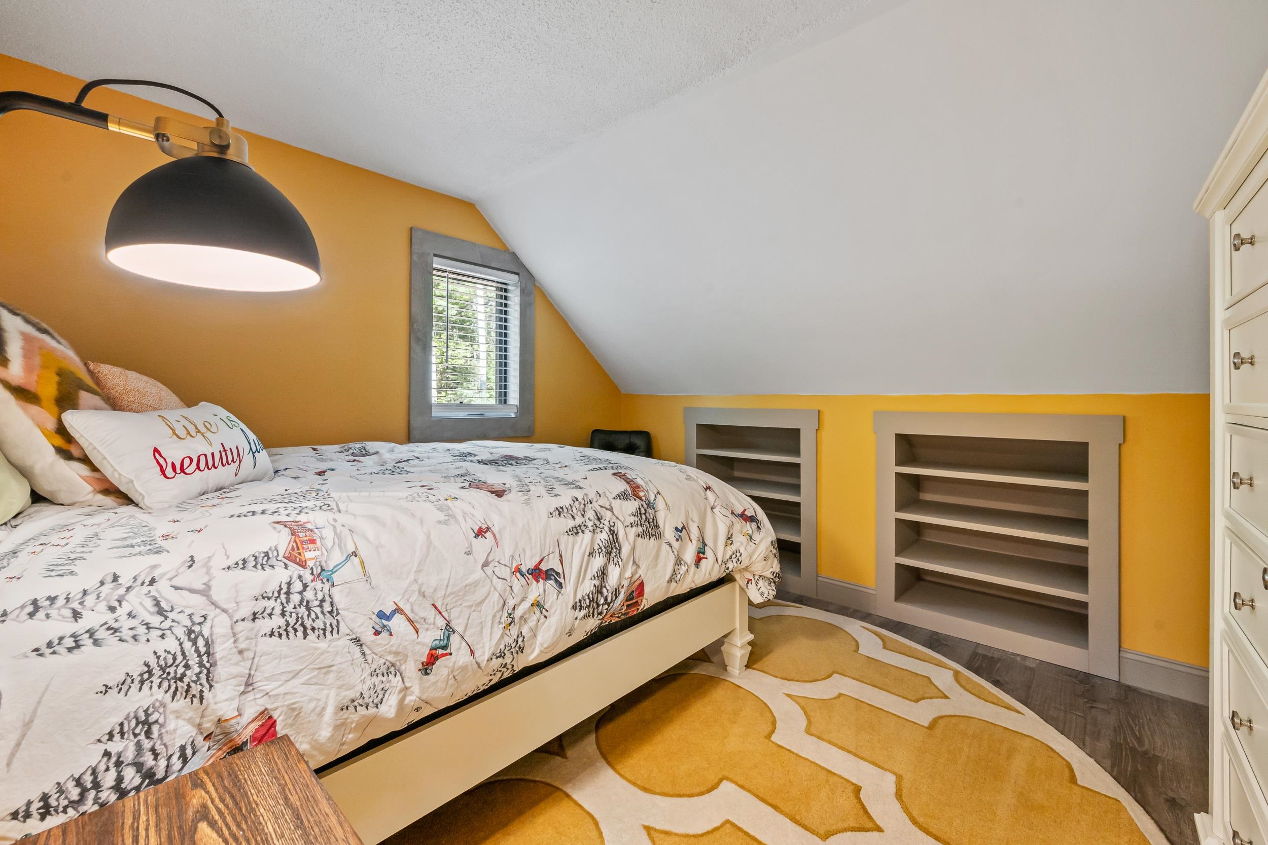 Attic bedroom with a ski-print bedspread, mustard walls, built-in shelves, and a geometric gold rug.