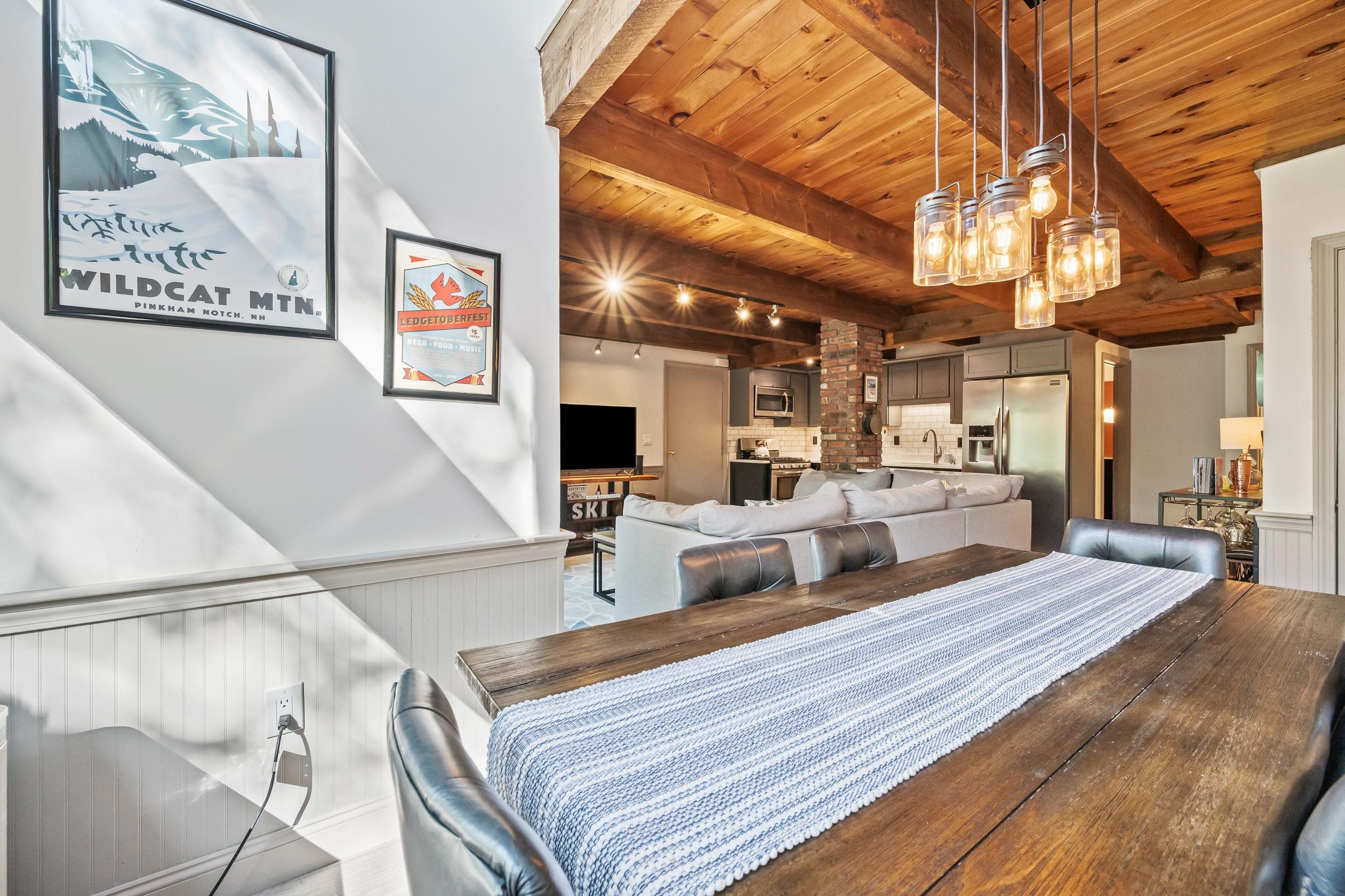Open-plan dining and living area with exposed timber beams, a rustic dining table, and a brick kitchen column.