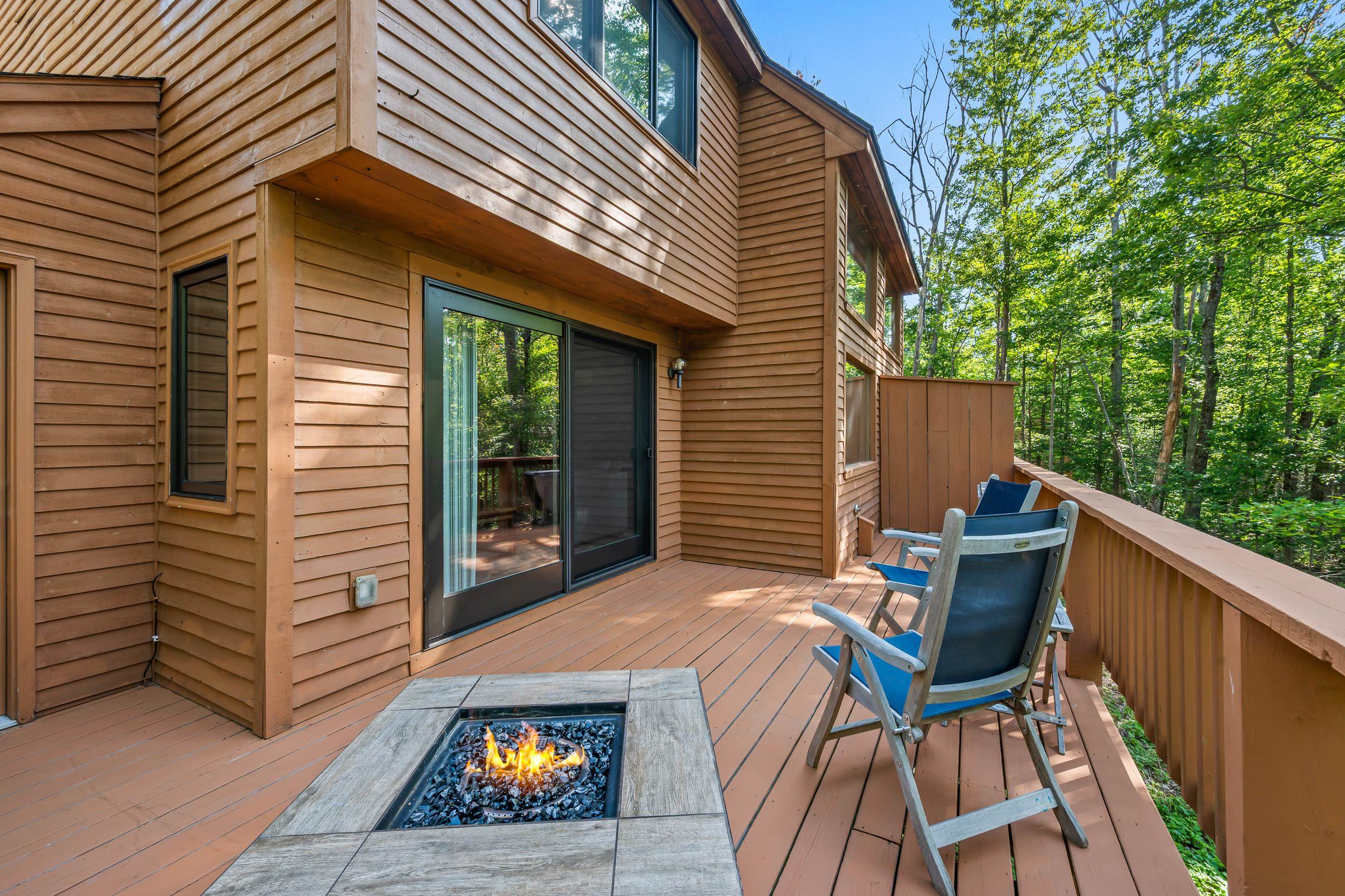 Wooden deck with outdoor chairs and a built-in fire pit beside a wood-sided home.