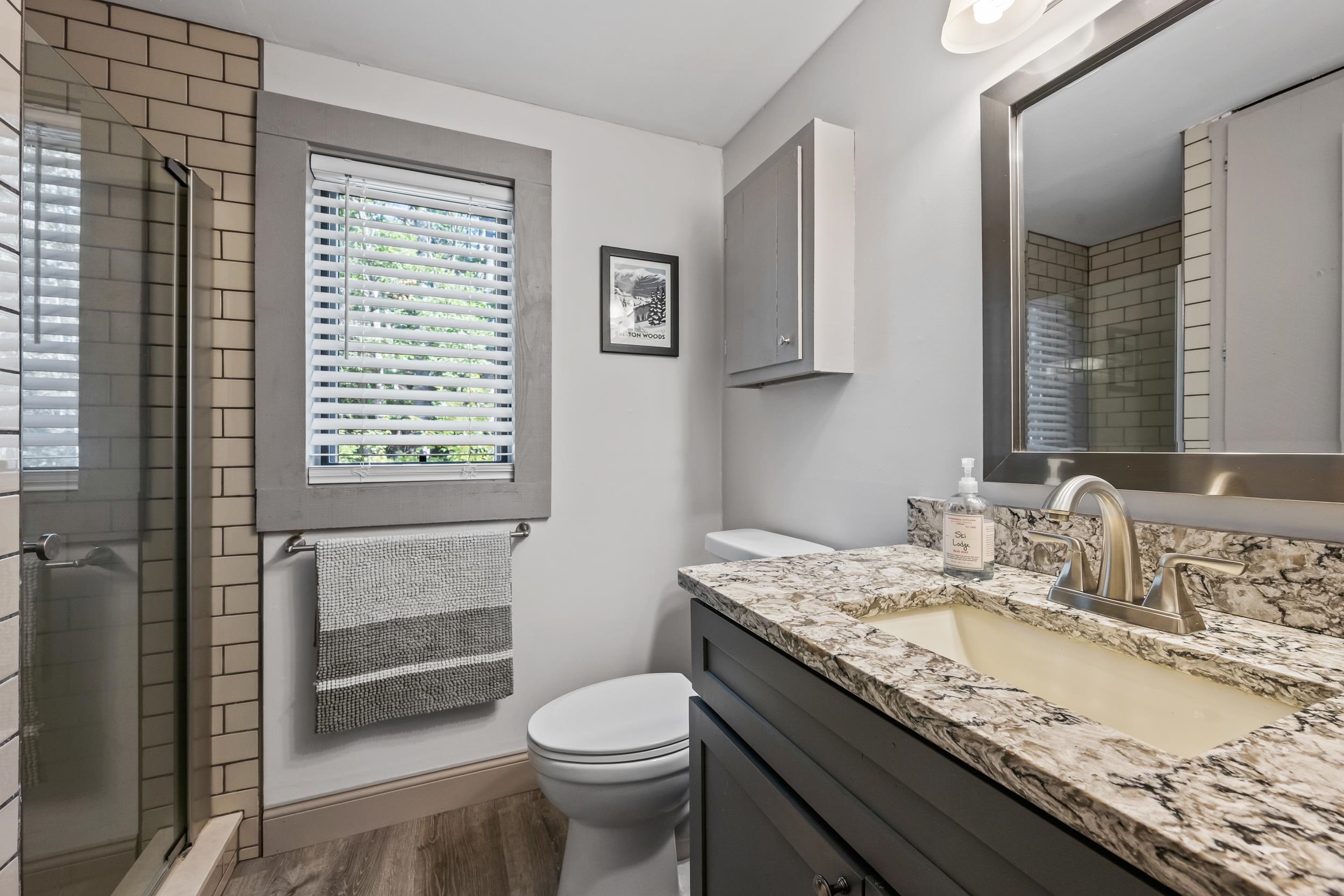 Bathroom with a glass shower, window with white blinds, and gray vanity with patterned stone countertop.
