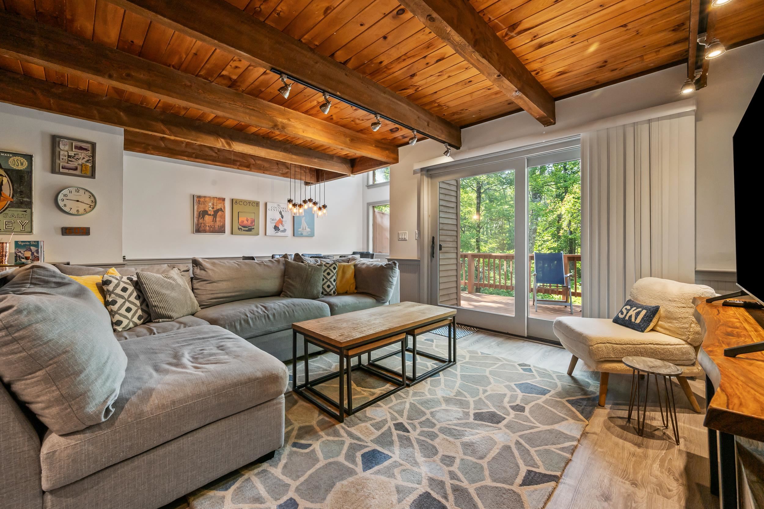 Living room with exposed timber ceiling, gray sectional, and sliding doors to a wooded deck.