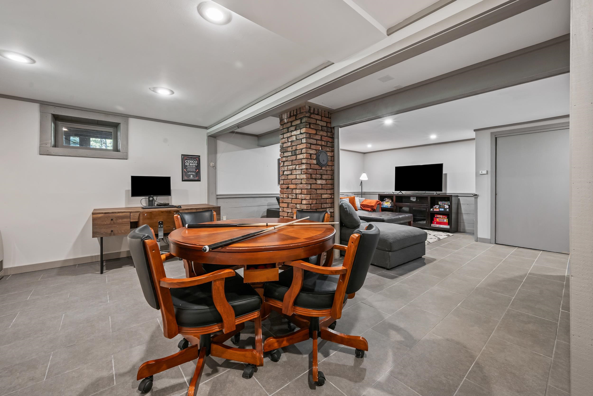 Recreation room with a round wooden game table, pool cues, exposed brick column, and adjoining TV lounge.