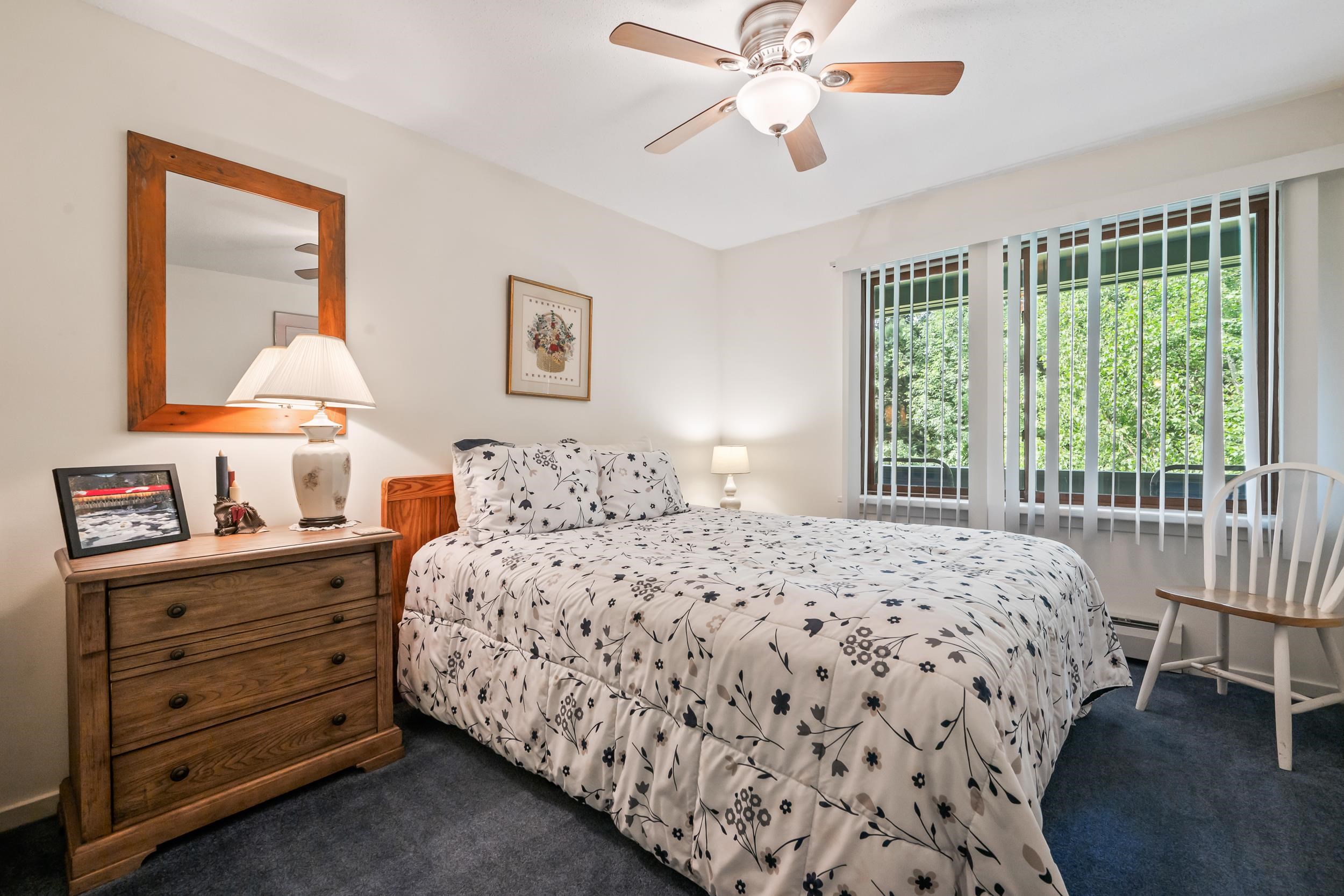 Bedroom with floral bedding, wooden dresser, ceiling fan, and a wide window overlooking greenery.