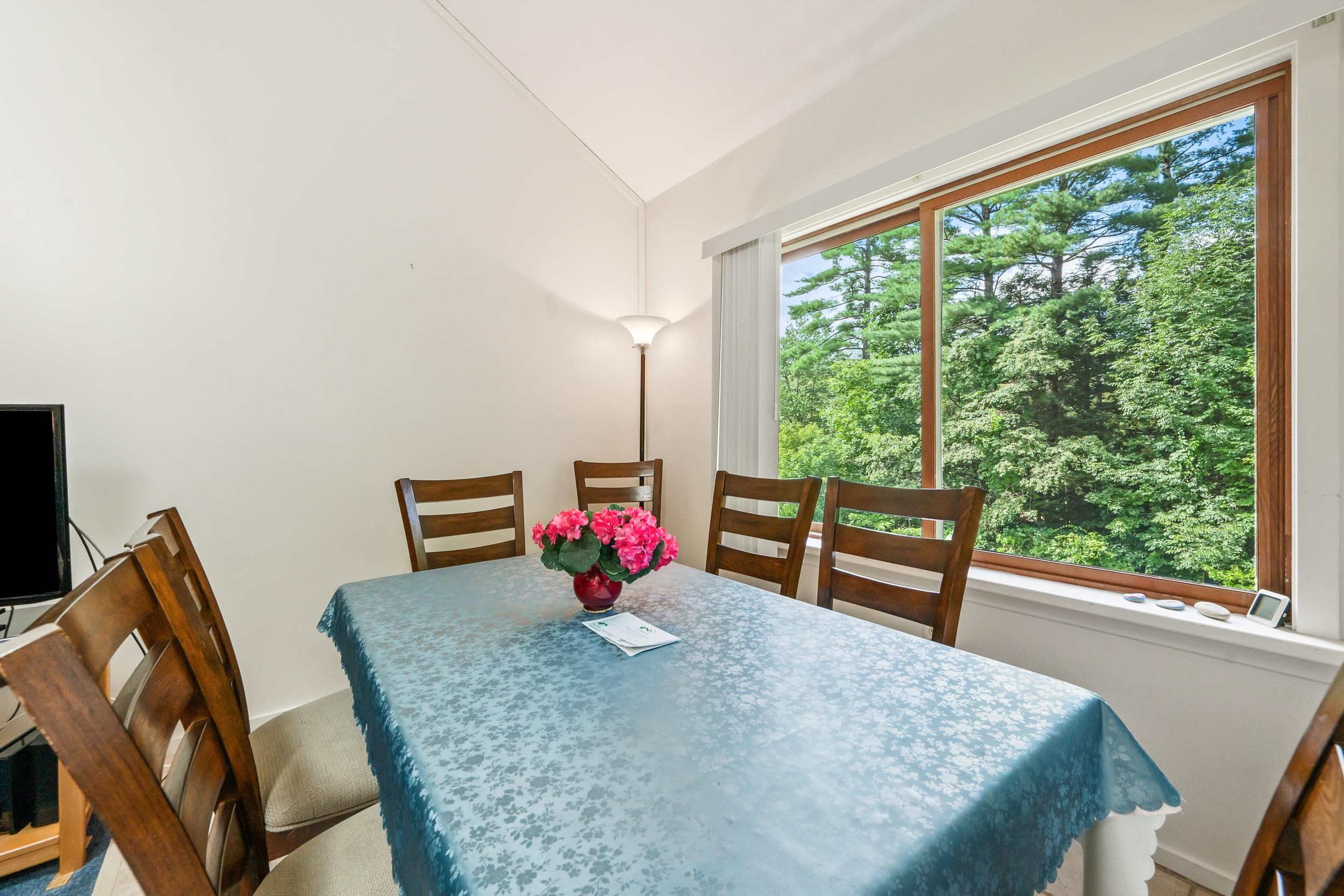 Dining area with a blue floral tablecloth, wooden chairs, and a large window overlooking trees.
