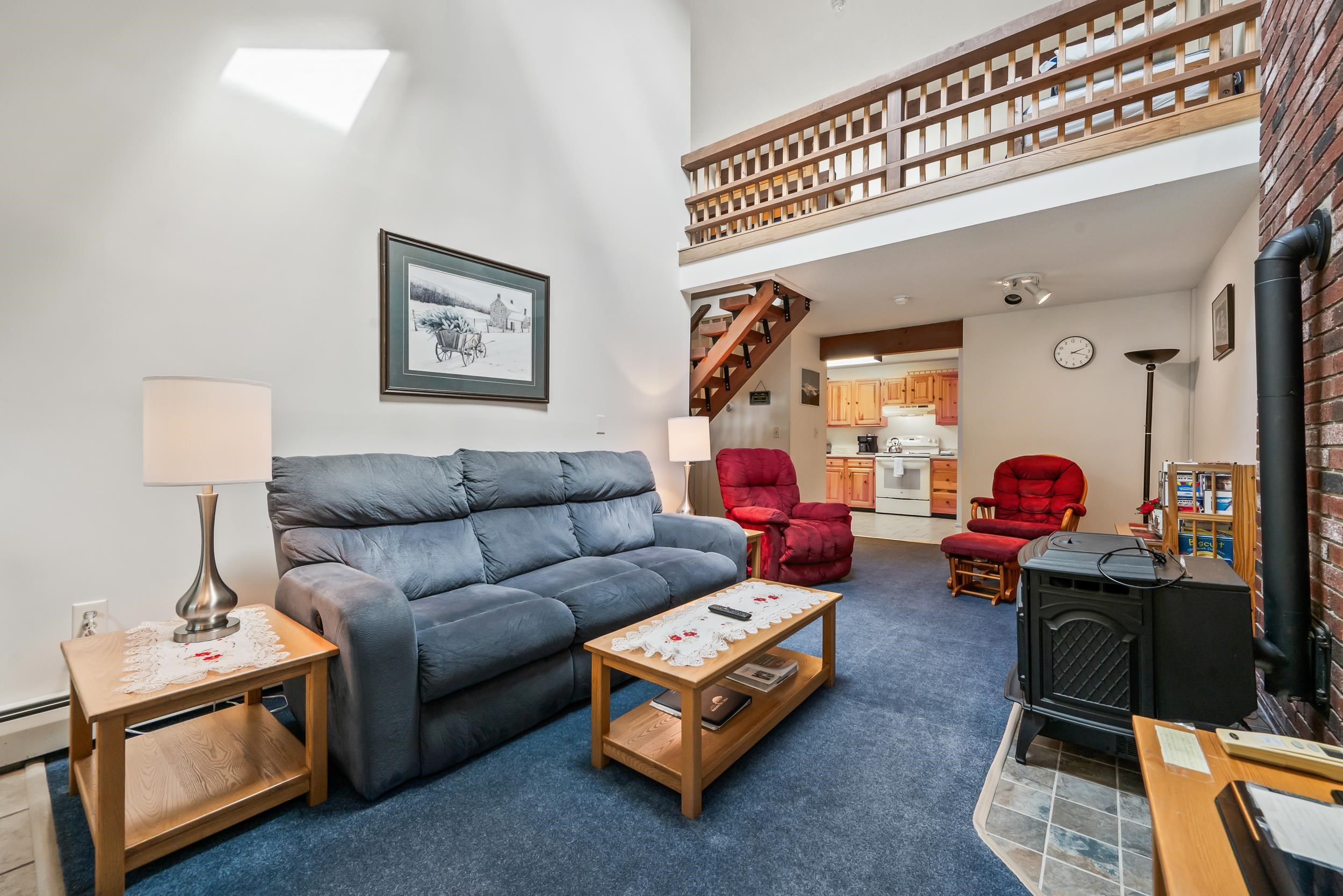 Living room with a blue sofa, red recliners, a black wood stove, and an open loft staircase.