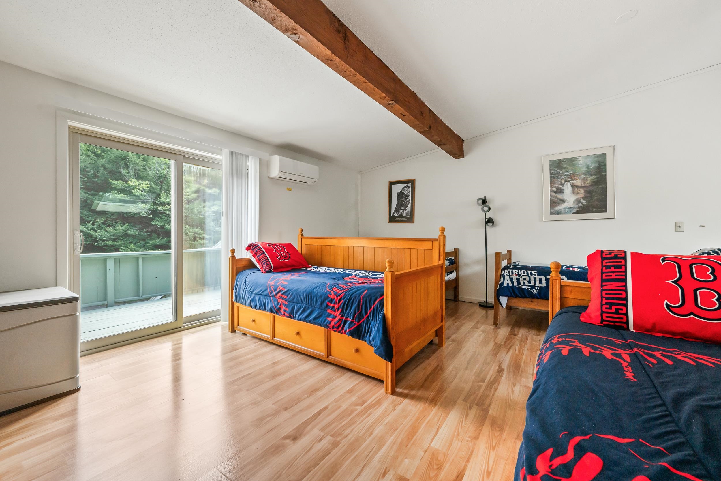 Bedroom with three wooden beds, sports-themed bedding, an exposed ceiling beam, and a wooded deck view.