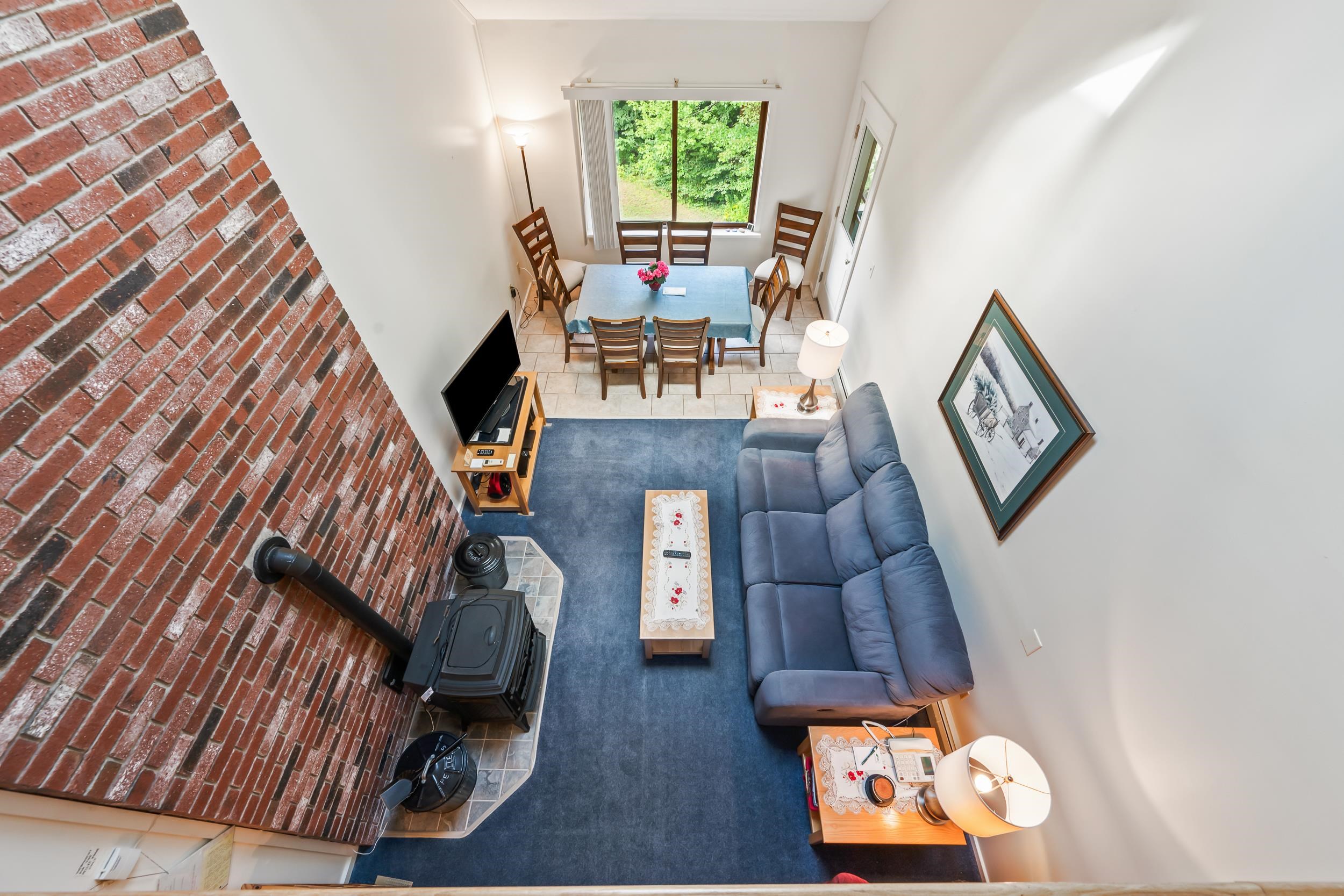Living and dining area with a brick wood-stove wall, blue sectional, and window overlooking greenery.