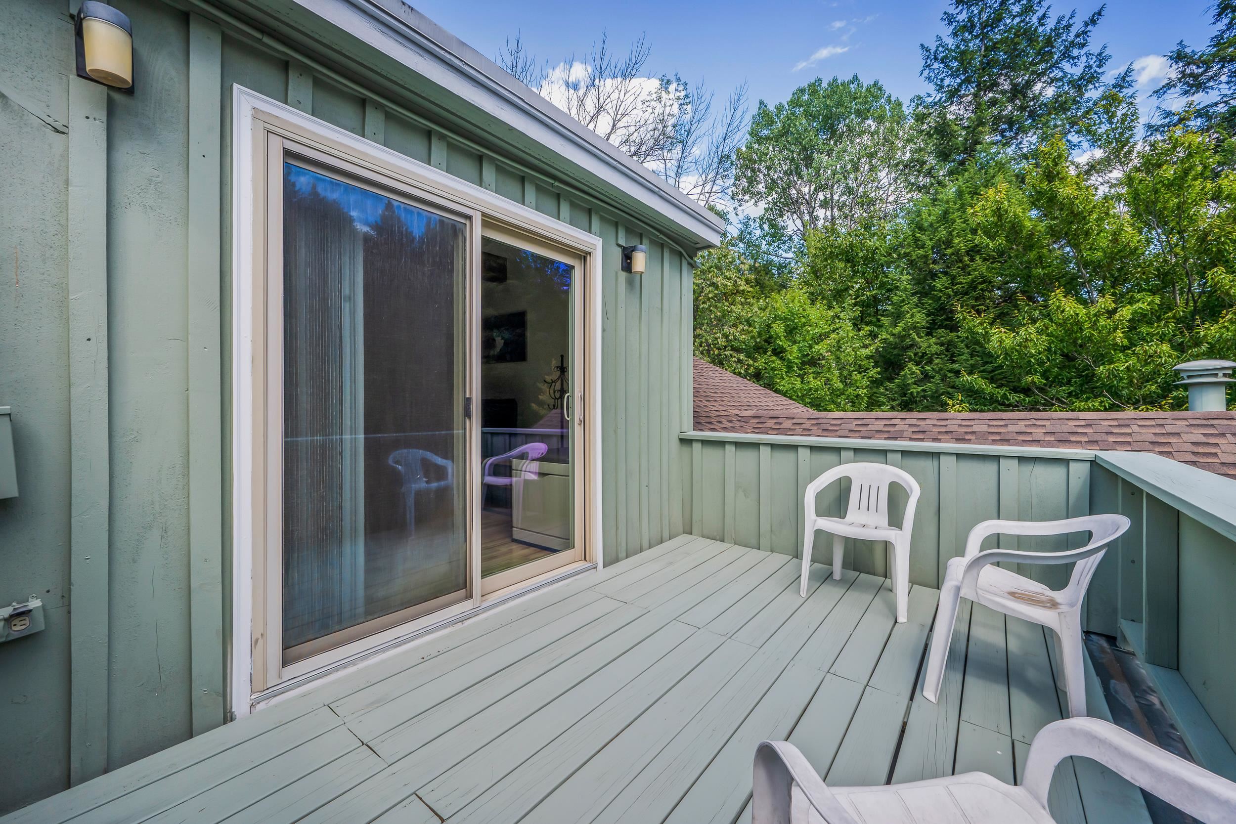 Raised deck with white chairs, sliding glass door, and leafy trees beyond the railing.