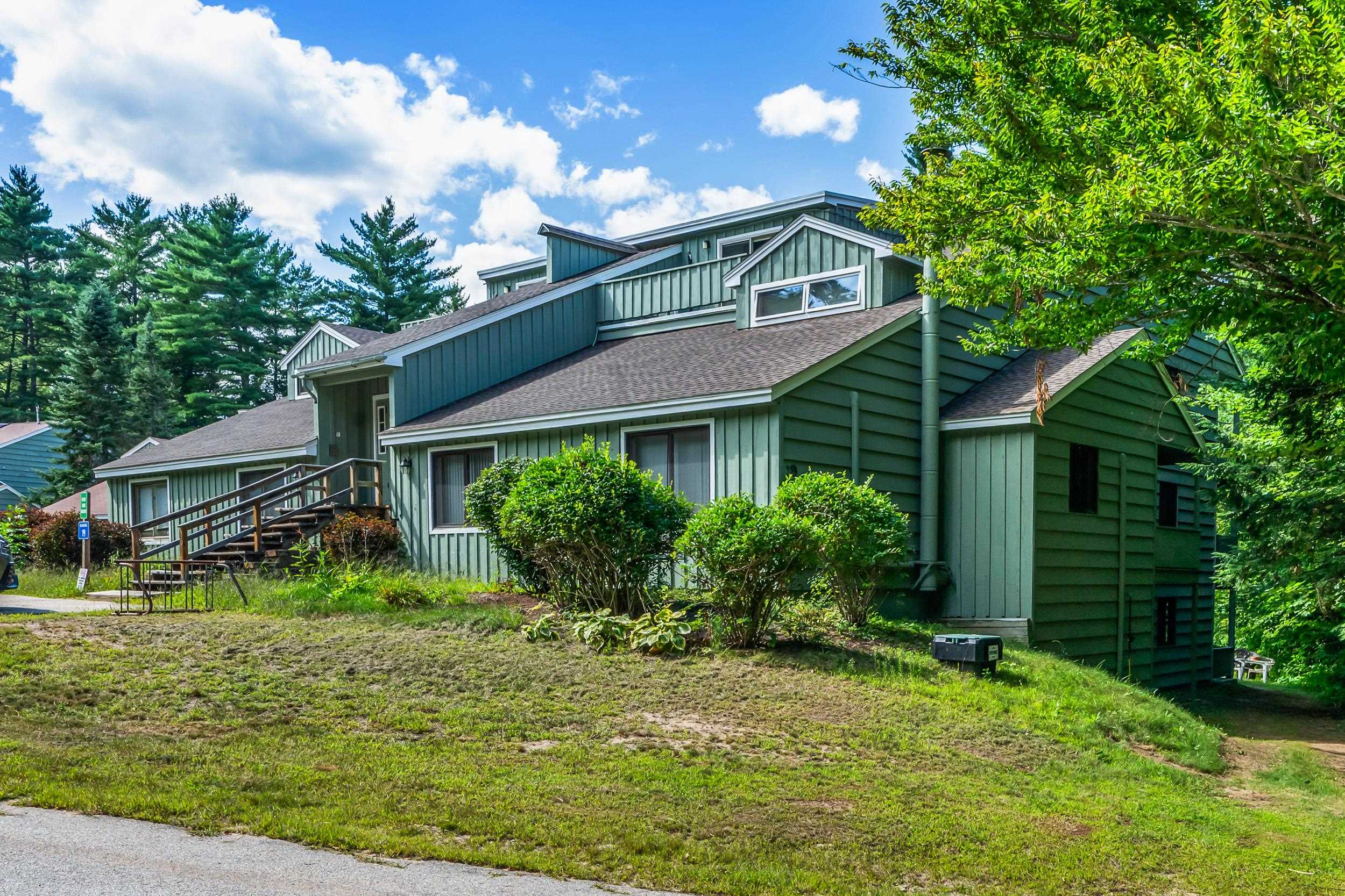 Green multi-level condominium with a wooden access ramp, front shrubs, and mature trees.