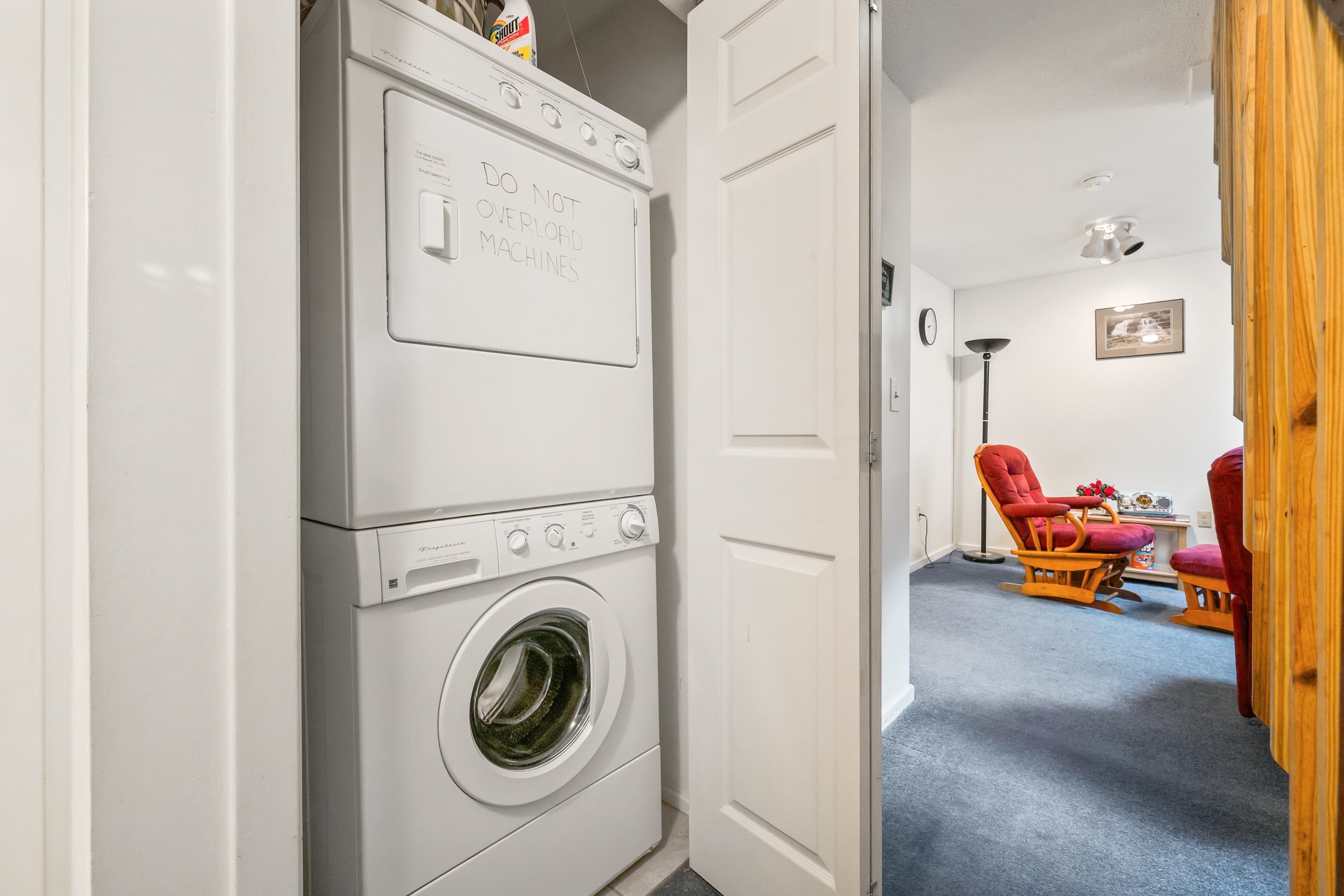 Stacked washer and dryer beside a sitting area with red-cushioned wooden chairs.
