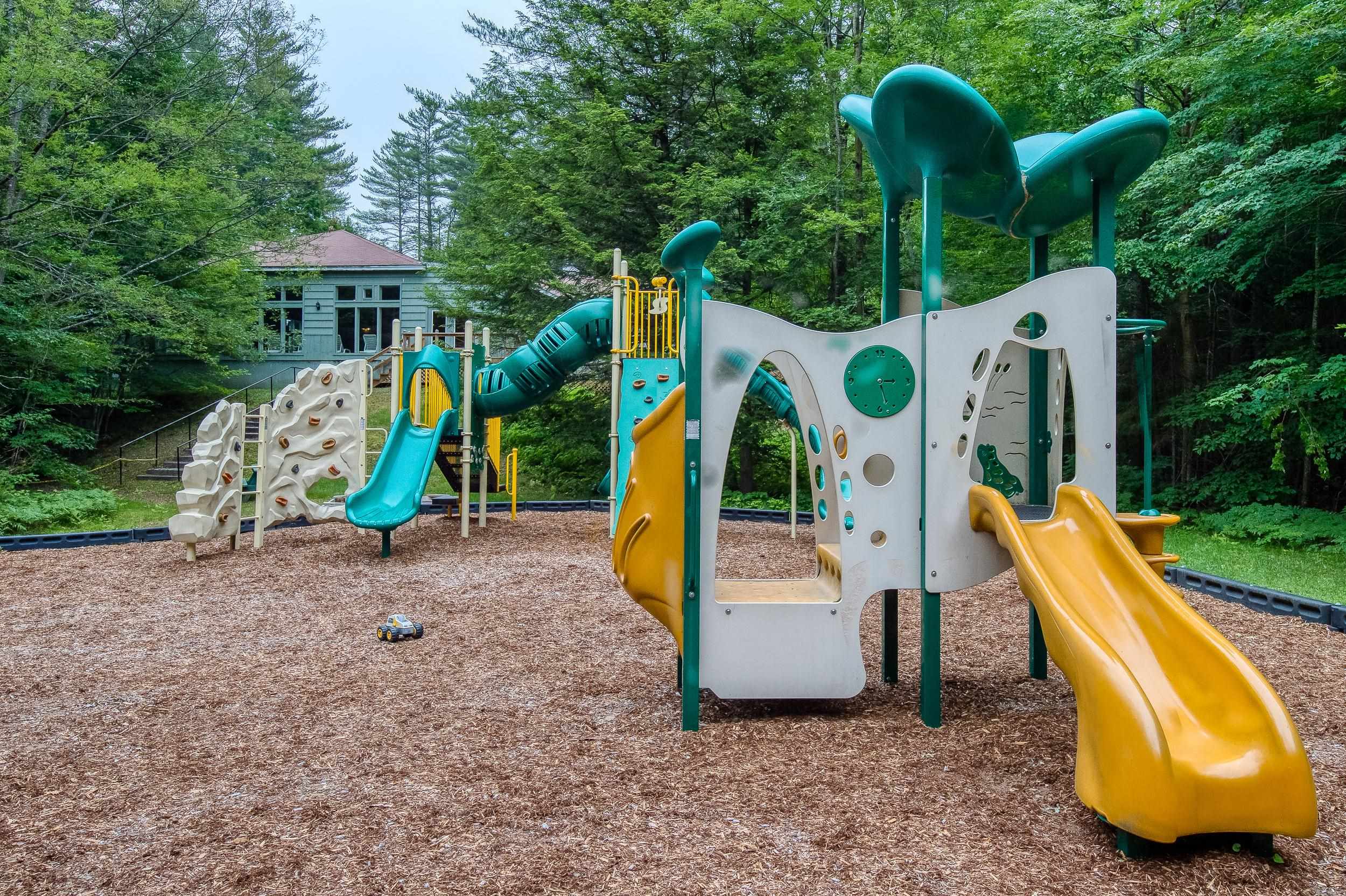 Playground with teal and yellow slides and climbing structures beside a wooded building.