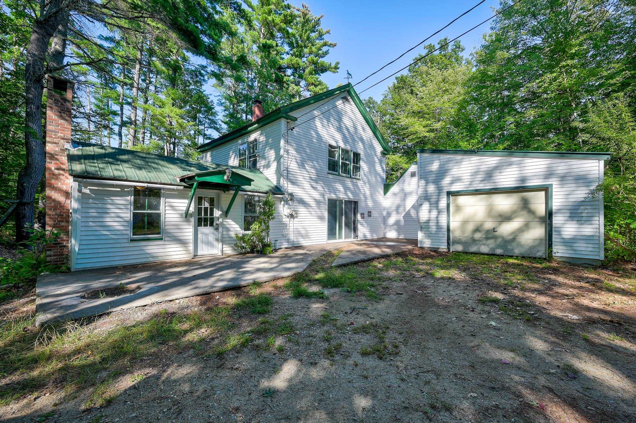 White two-story house with green metal roofs, attached garage, and brick chimney among mature trees.