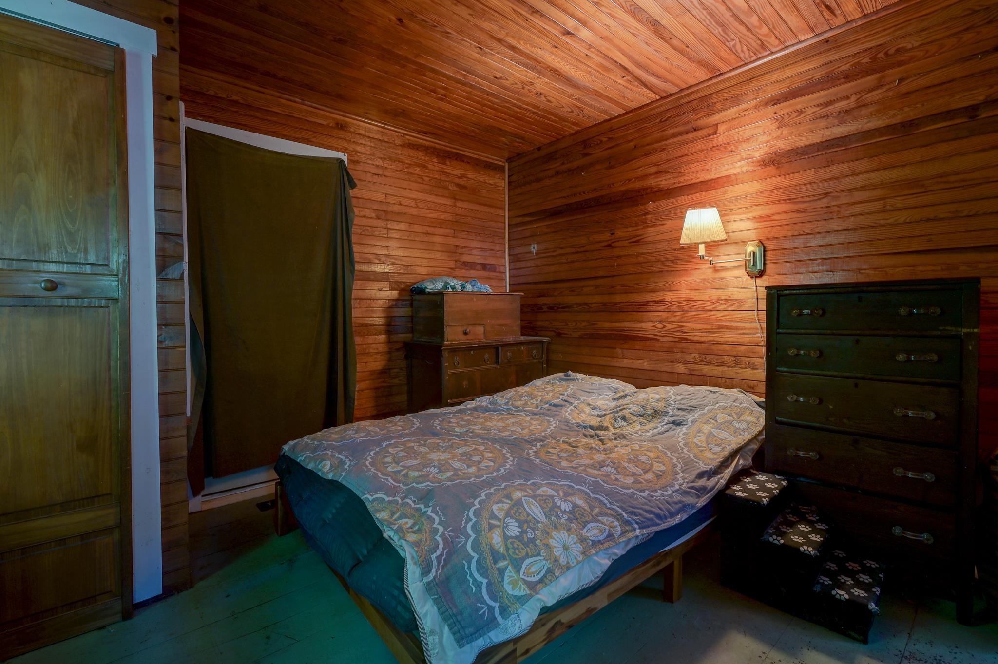 Wood-paneled bedroom with a patterned bedspread, two dressers, and a lit wall sconce.