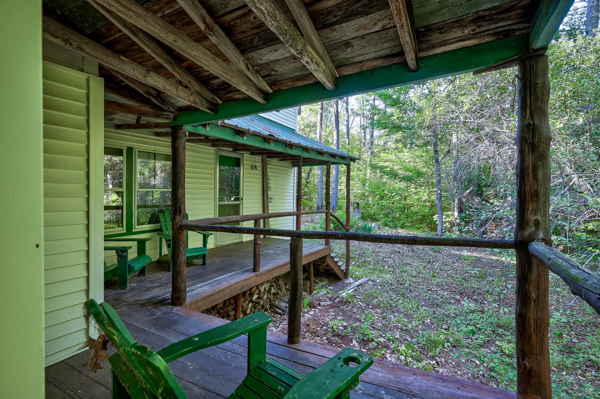 Covered wooden porch with green chairs and rustic beams overlooking a wooded yard.