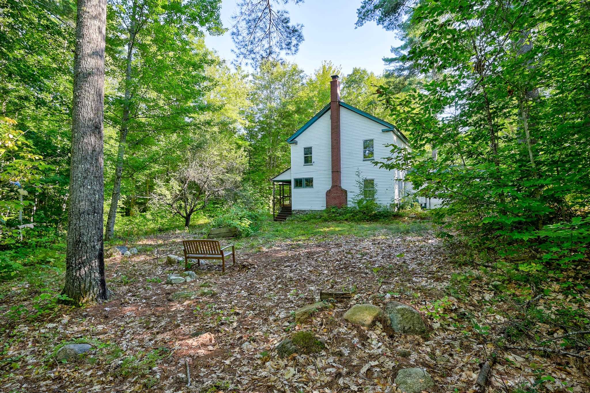 White two-story house with a tall brick chimney in a wooded yard with a wooden bench.