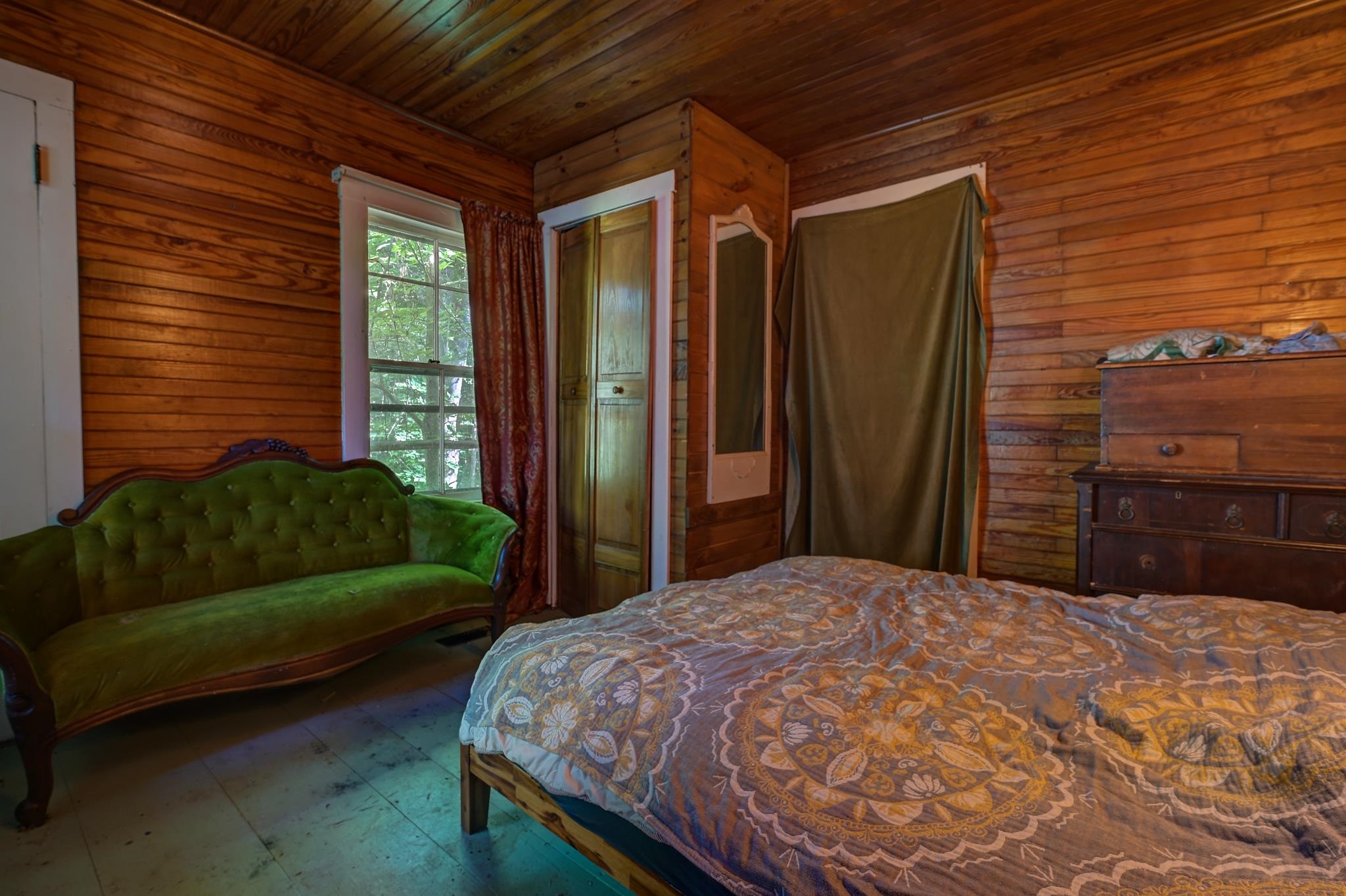 Wood-paneled bedroom with a patterned bedspread, green velvet settee, and leafy views through the window.