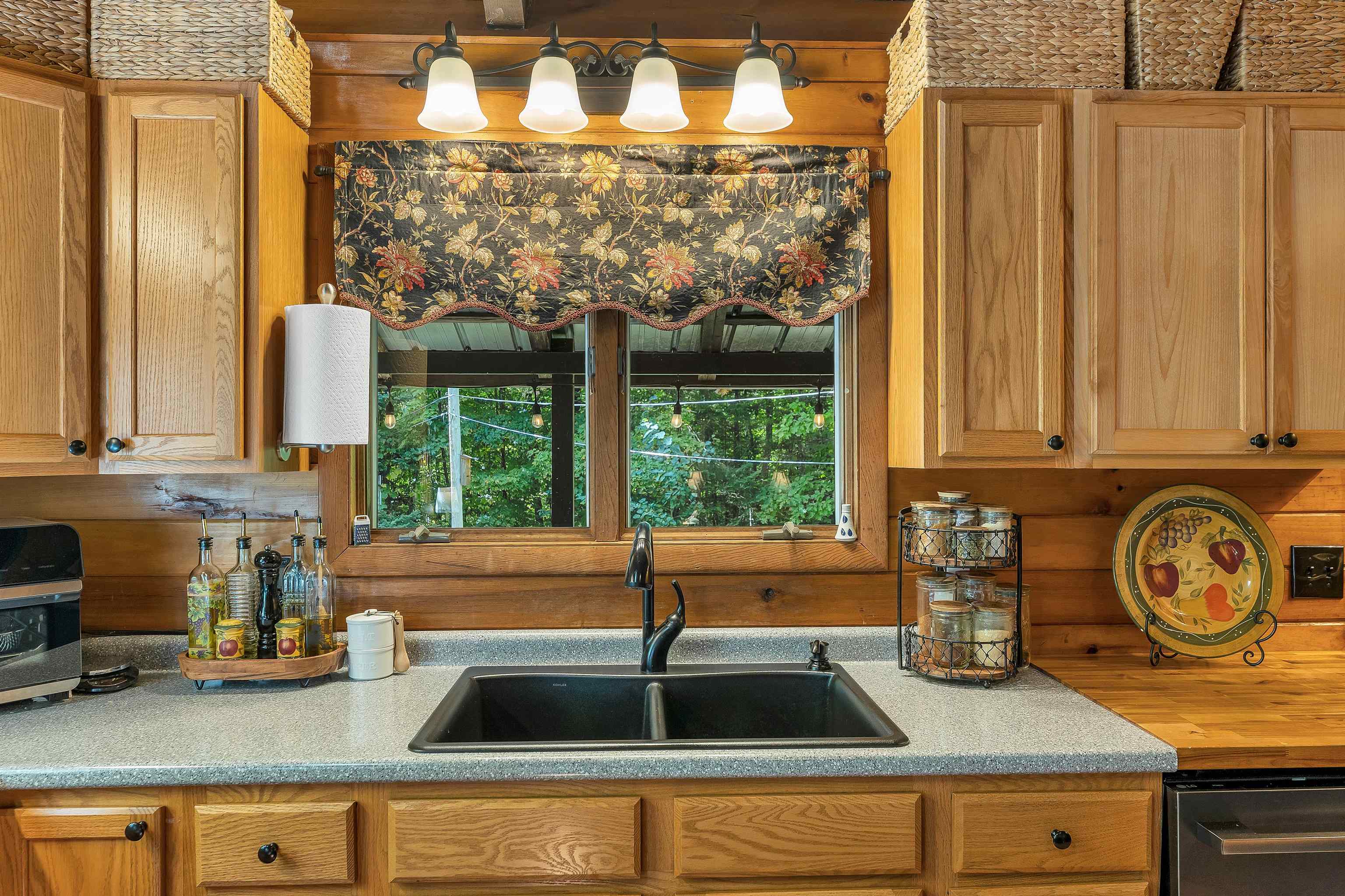 Rustic kitchen sink beneath a window, surrounded by honey-toned wood cabinets and a floral valance.