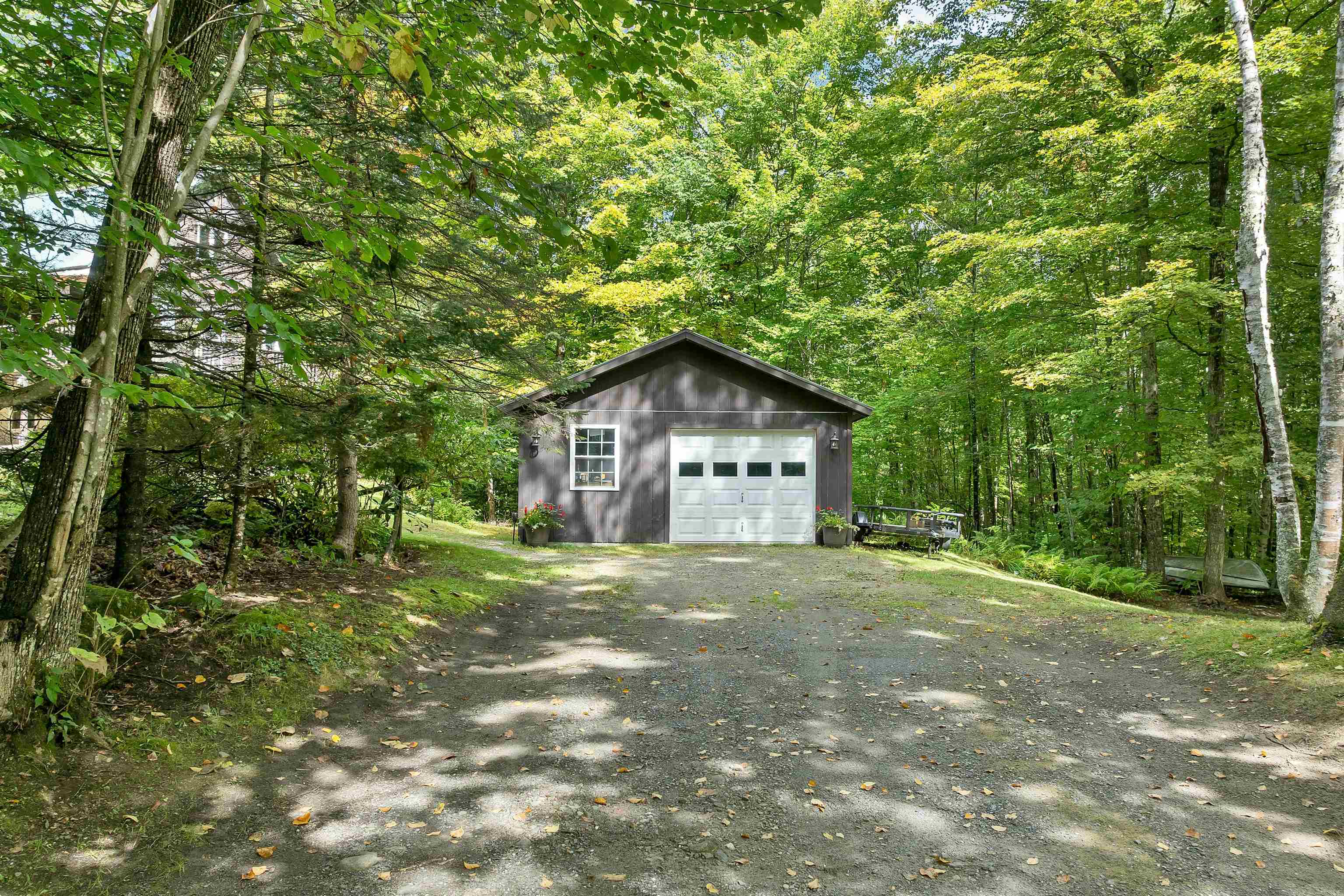 Detached gray garage with a white overhead door, surrounded by trees beside a gravel driveway.