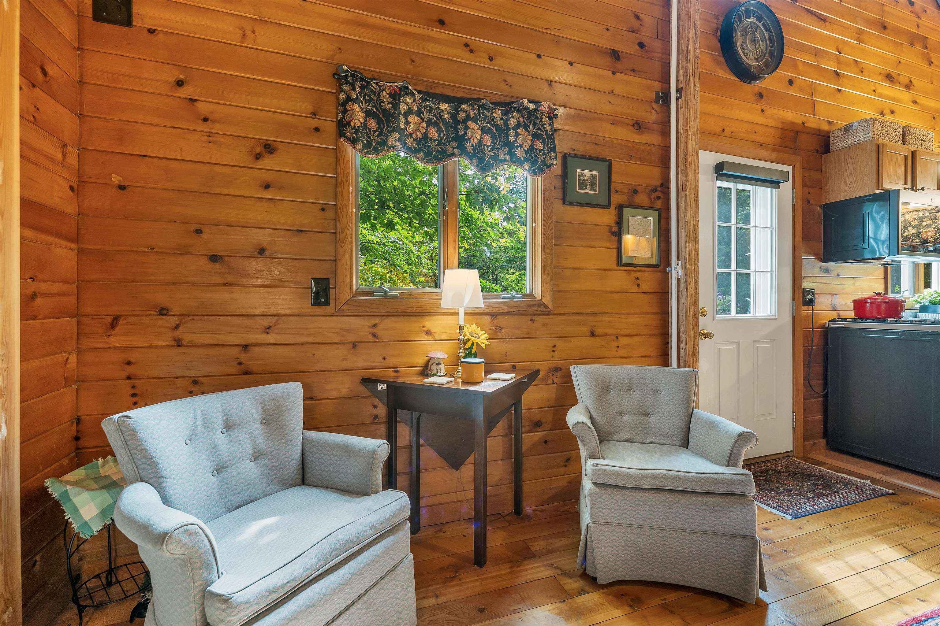 Cabin living area with pale armchairs, knotty-pine walls, and a window overlooking leafy trees.