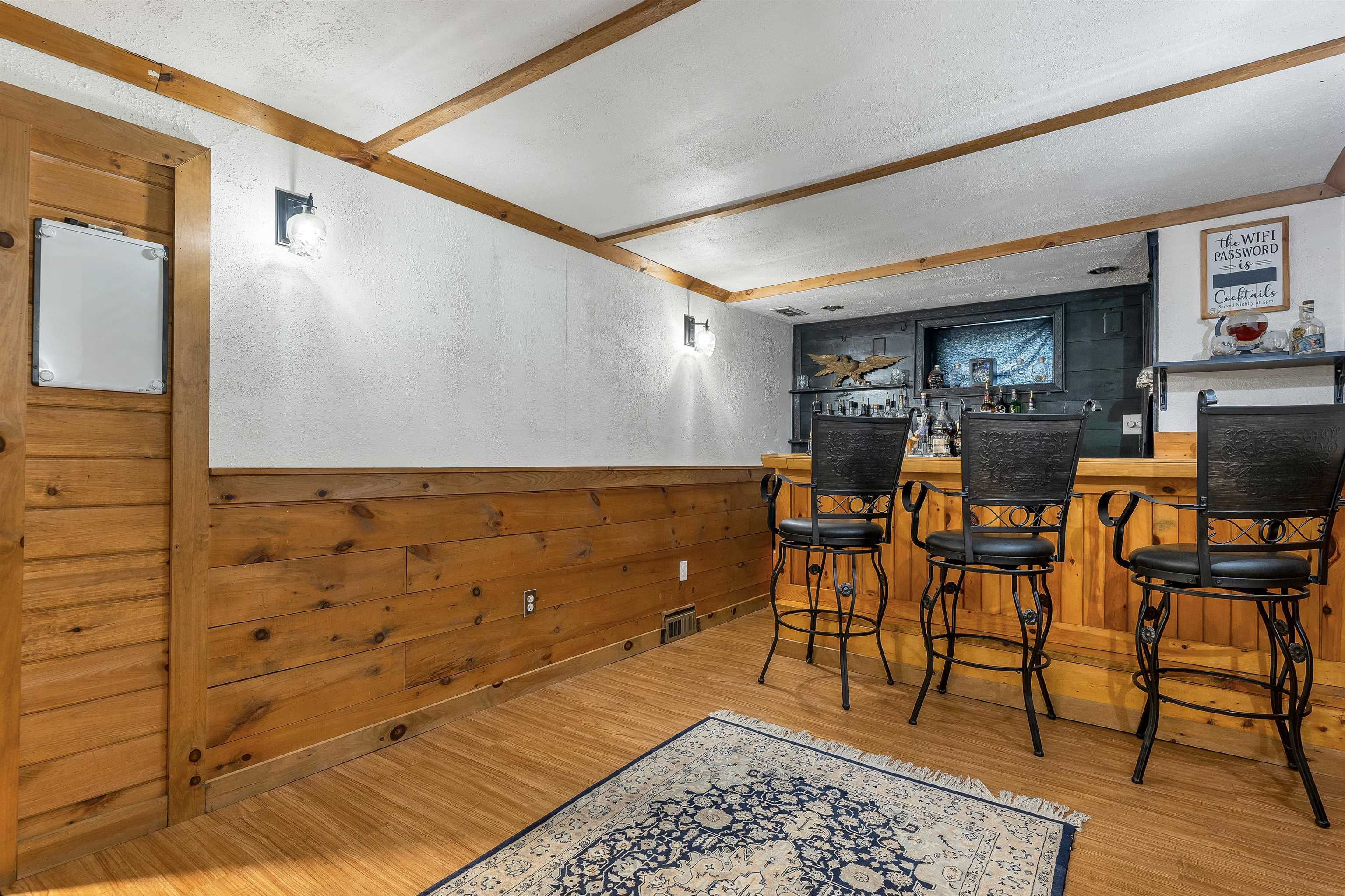 Wood-paneled bar with three black stools, exposed beams, and a patterned rug.