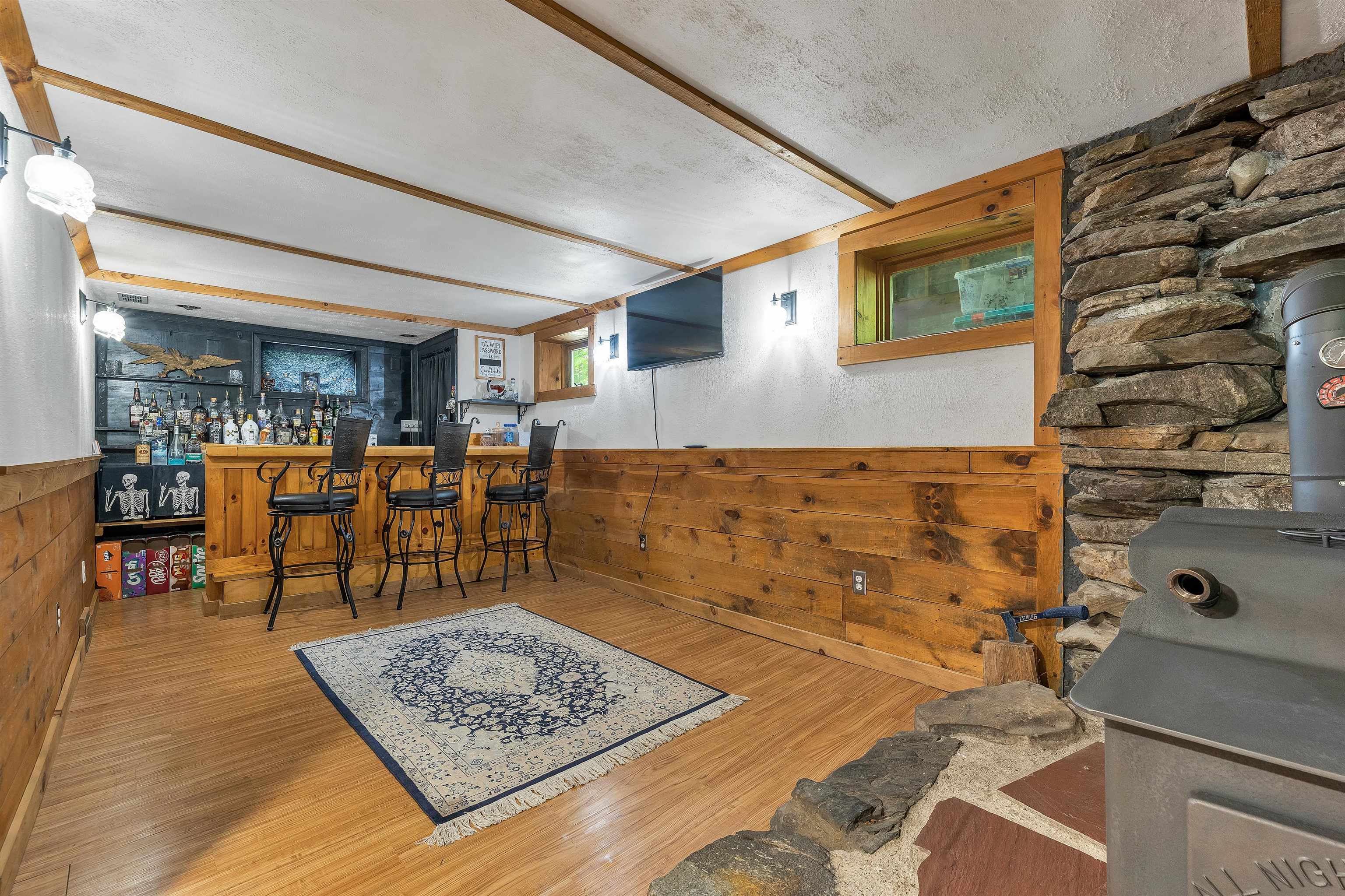 Recreation room with a wooden bar and stools beside a stone hearth and wood stove.