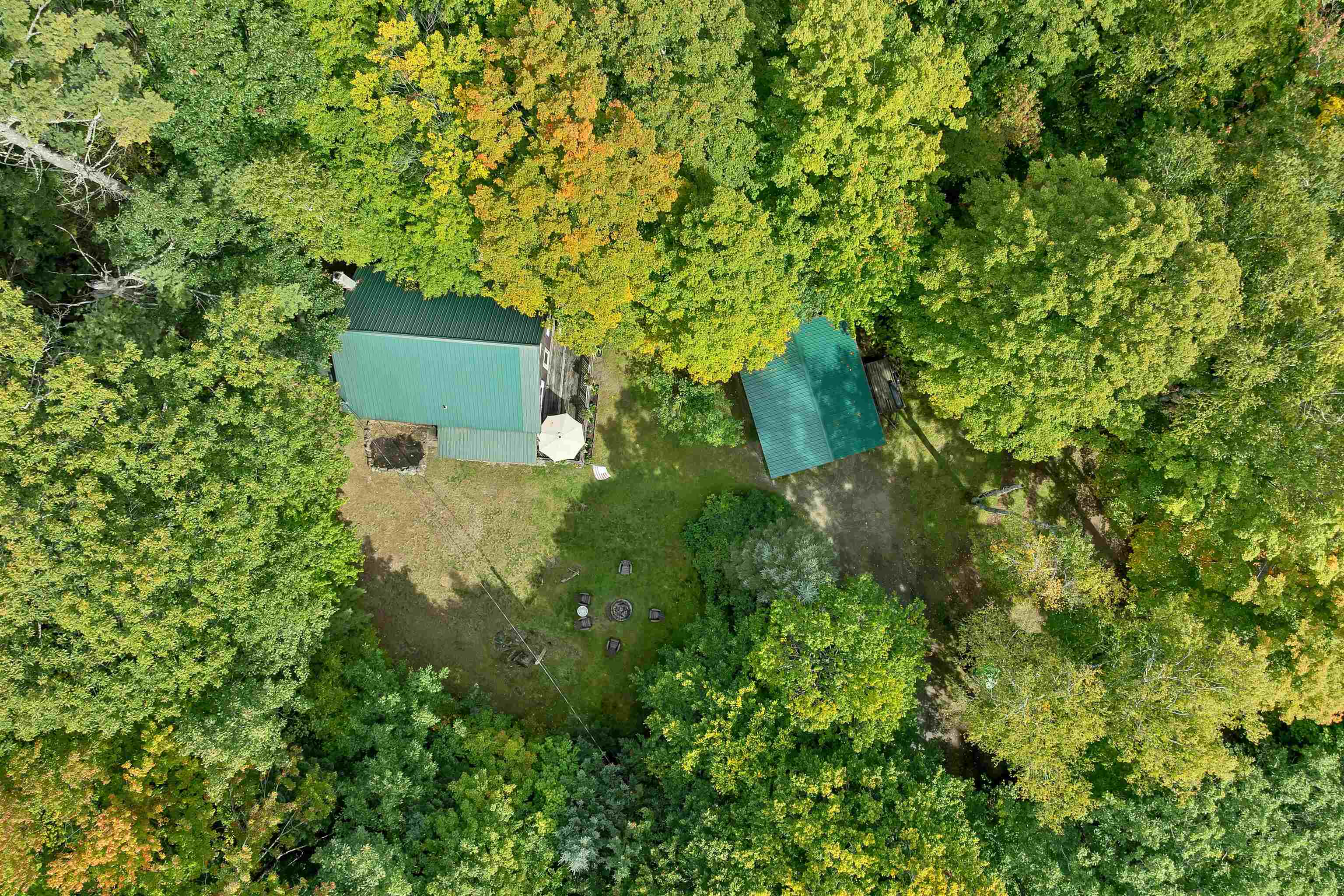 Aerial view of a wooded property with a green-roofed house, outbuilding, lawn, and fire pit.