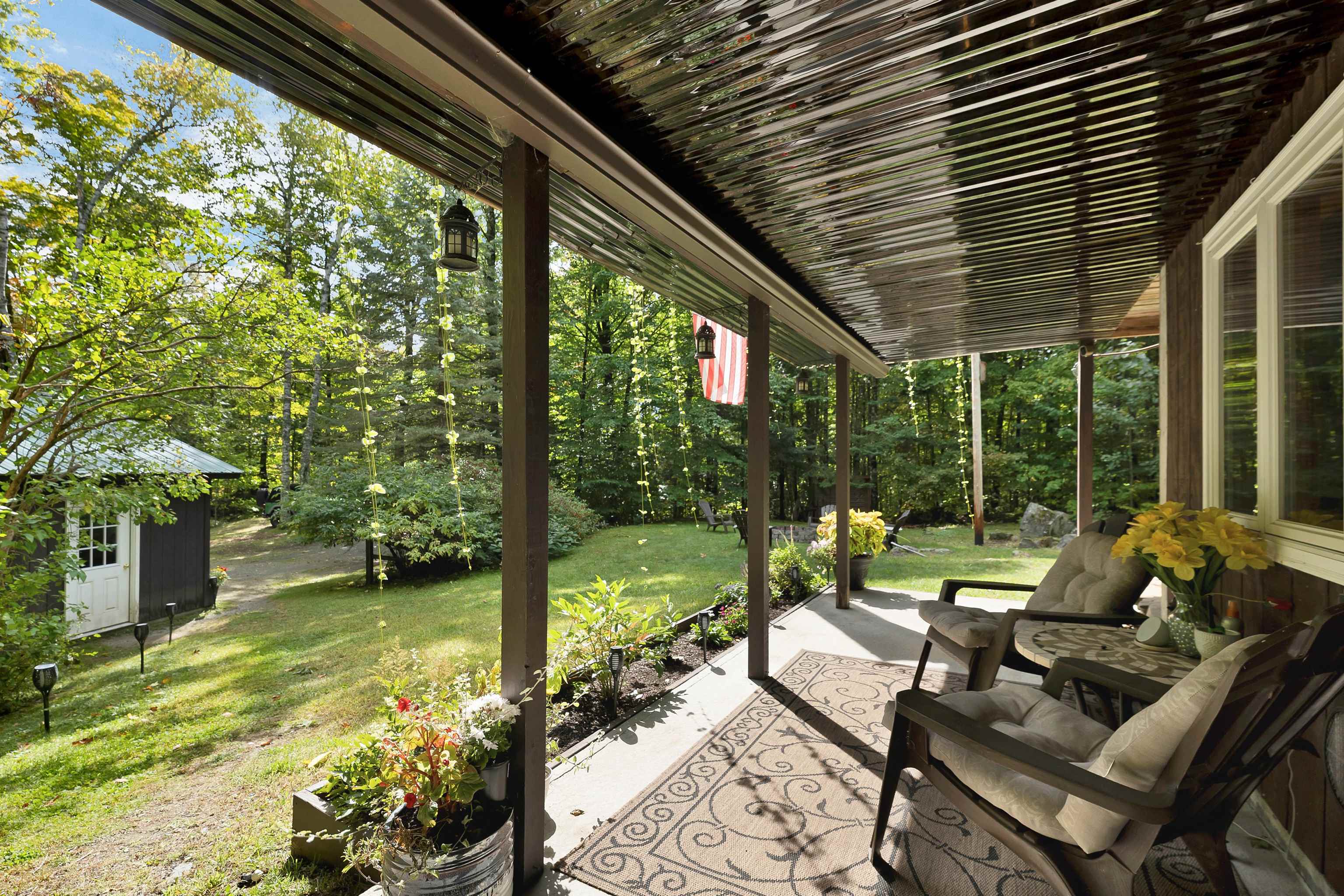Covered porch with cushioned chairs and potted flowers overlooking a leafy yard.