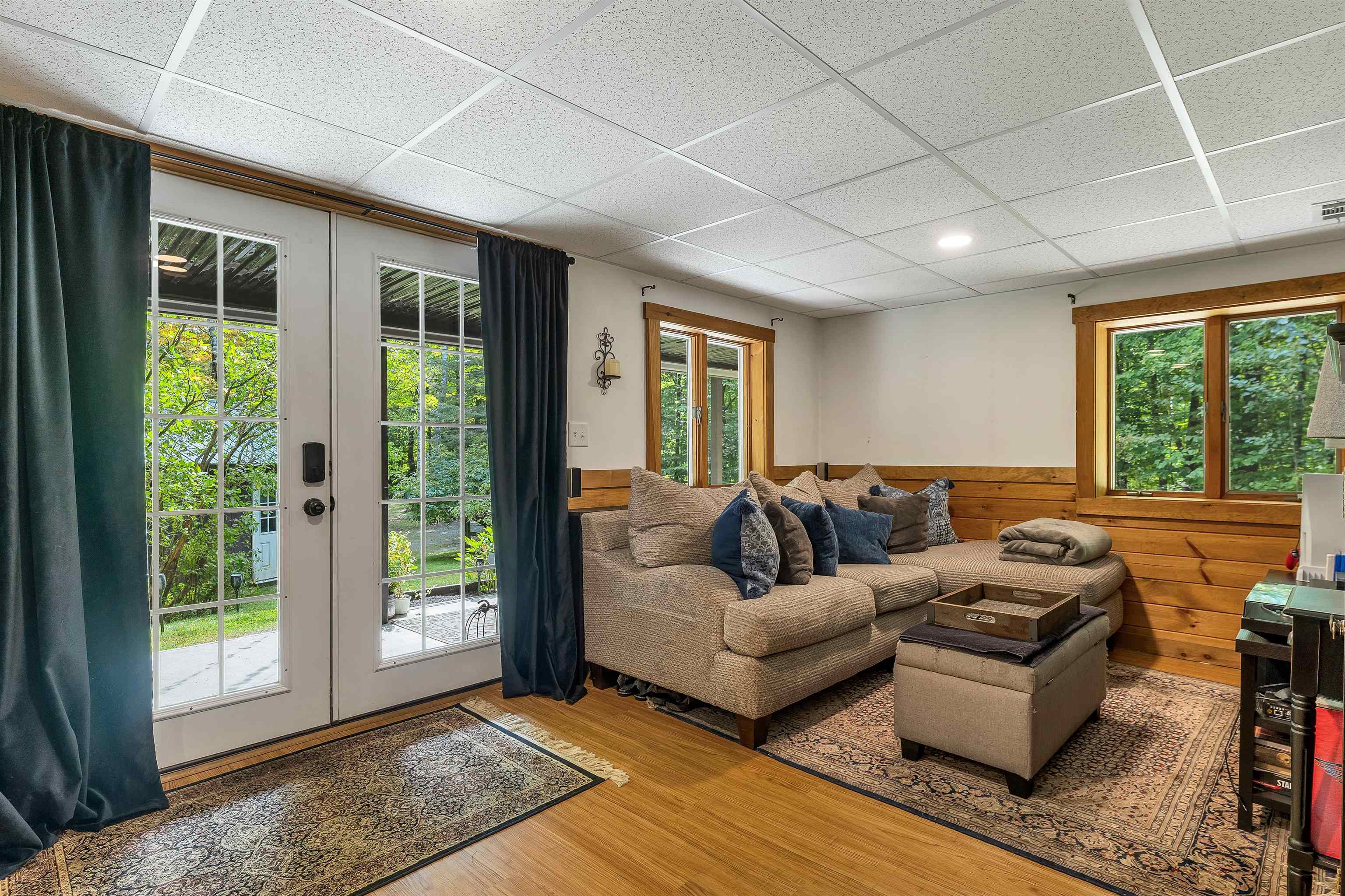 Furnished living room with a sectional sofa, wood-paneled walls, and glass doors opening toward greenery.
