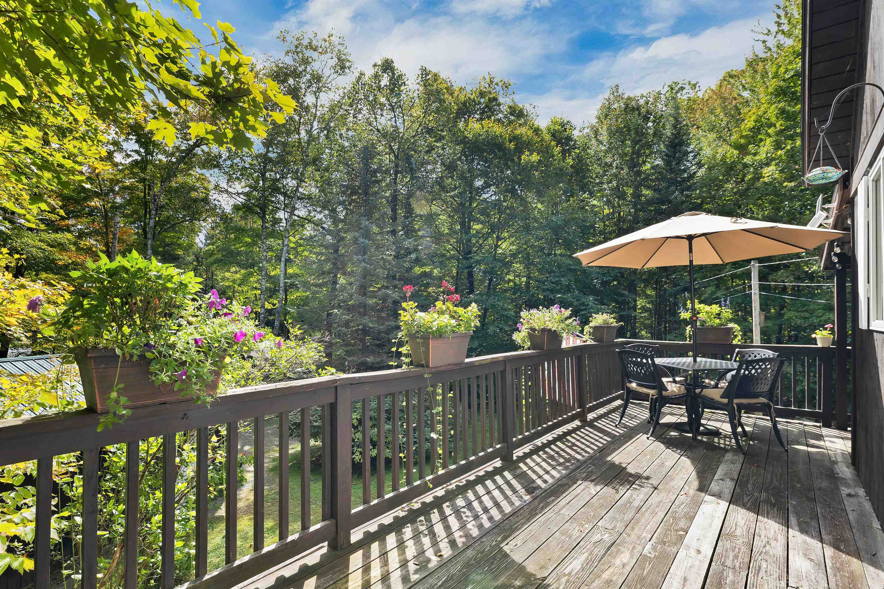 Wooden deck with outdoor dining set, umbrella, flower planters, and wooded surroundings