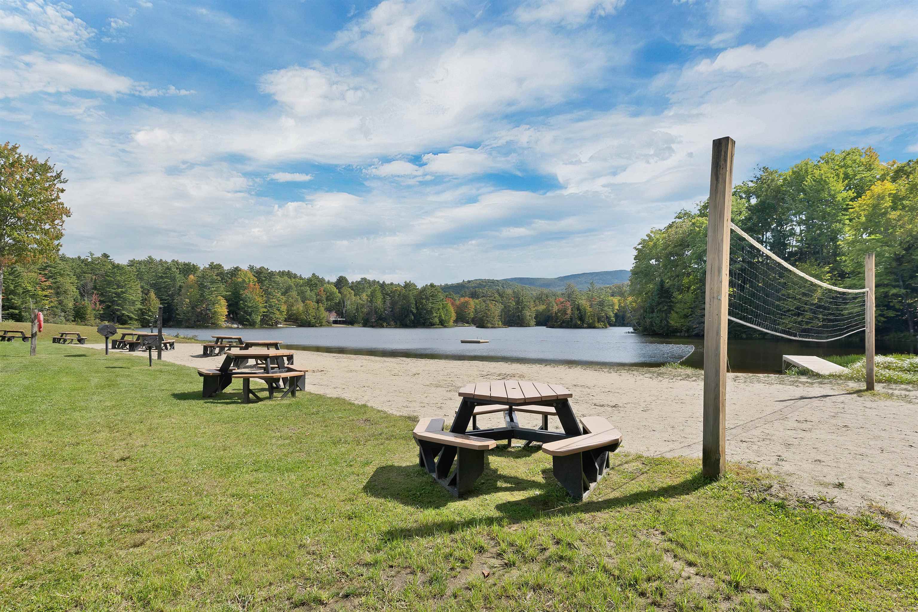 Lakeside beach with picnic tables and a volleyball net beside a wooded lake.