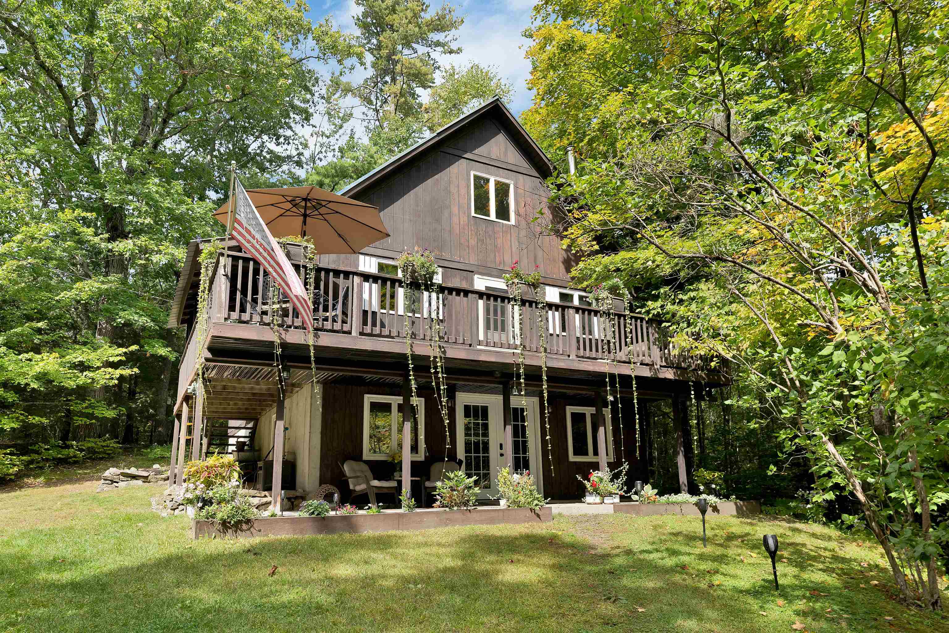 Wooded two-story home with a raised deck, American flag, tan umbrella, and covered patio.