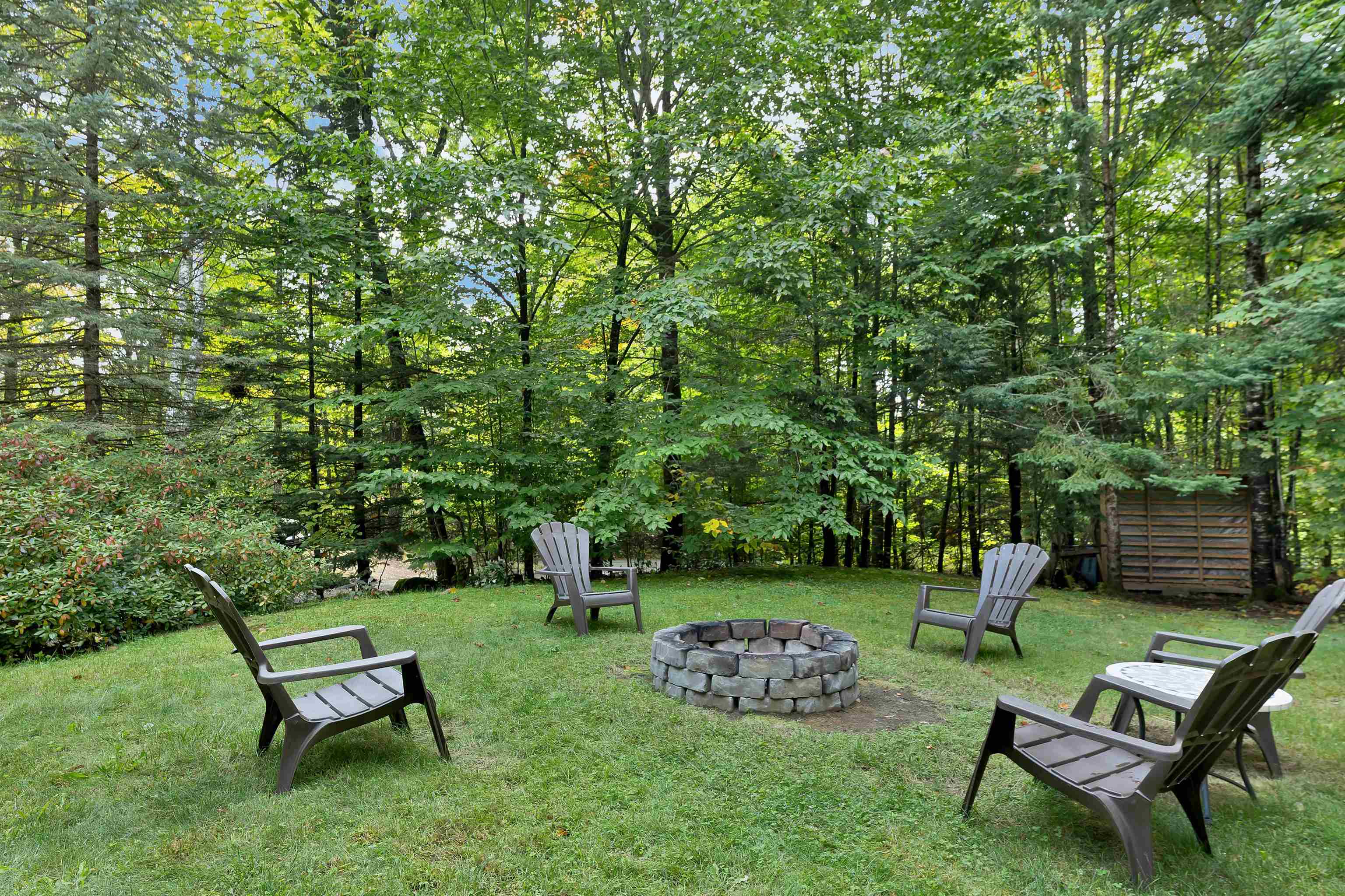 Wooded backyard with Adirondack chairs around a circular stone fire pit.
