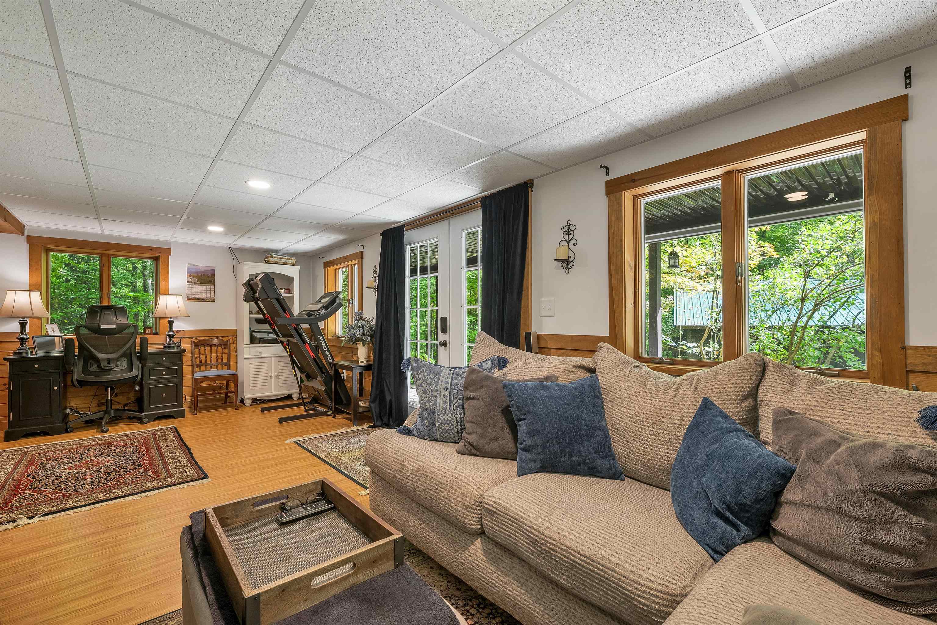 Living room with a beige sectional, wood-trimmed windows, and an office desk and treadmill.