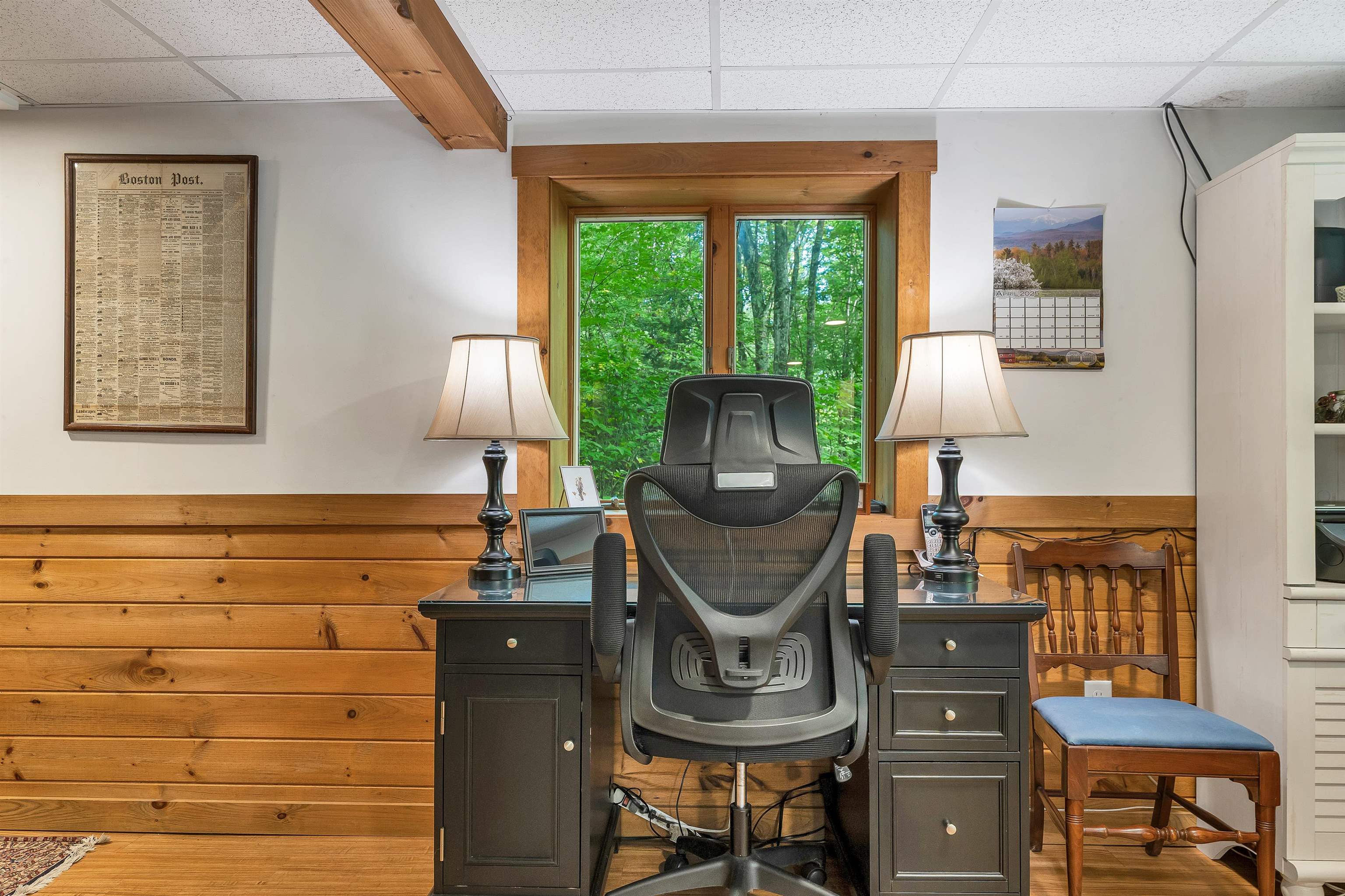 Home office with a dark desk and ergonomic chair, wood paneling, and a window overlooking leafy trees.