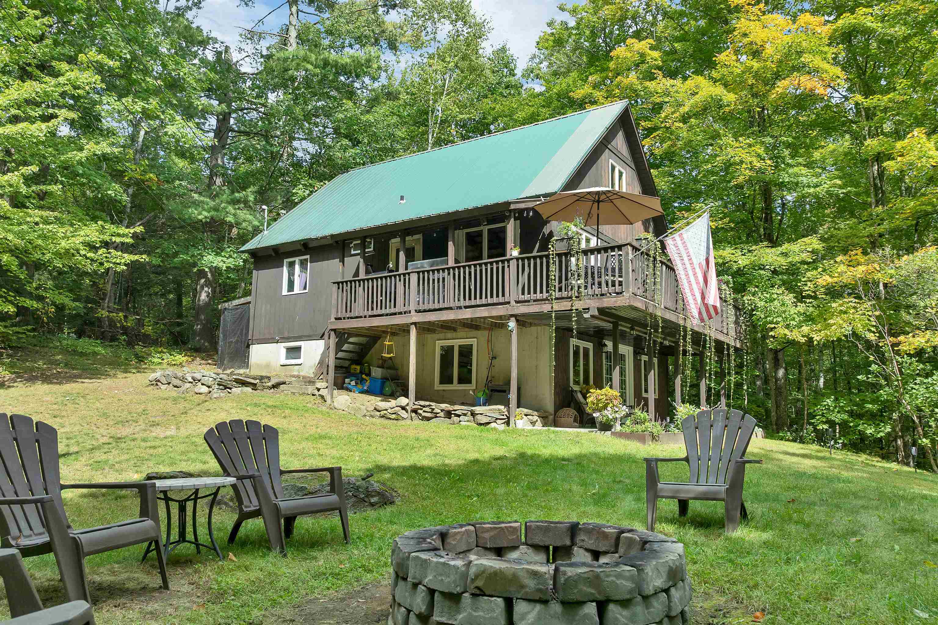 Wooded two-story home with a green metal roof, elevated deck, Adirondack chairs, and stone fire pit.