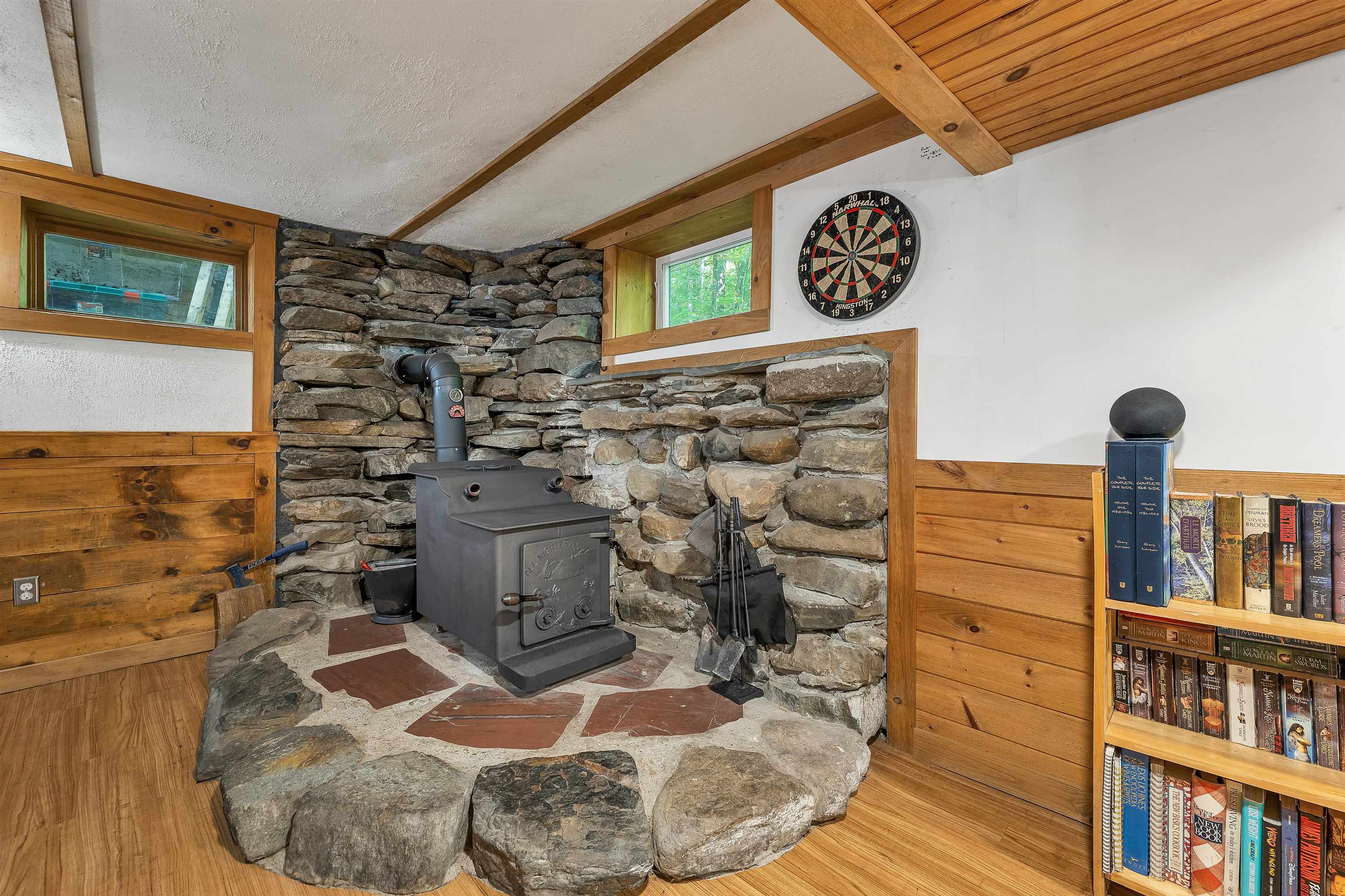 Black wood stove on a broad stone hearth, with exposed beams and a bookcase nearby.
