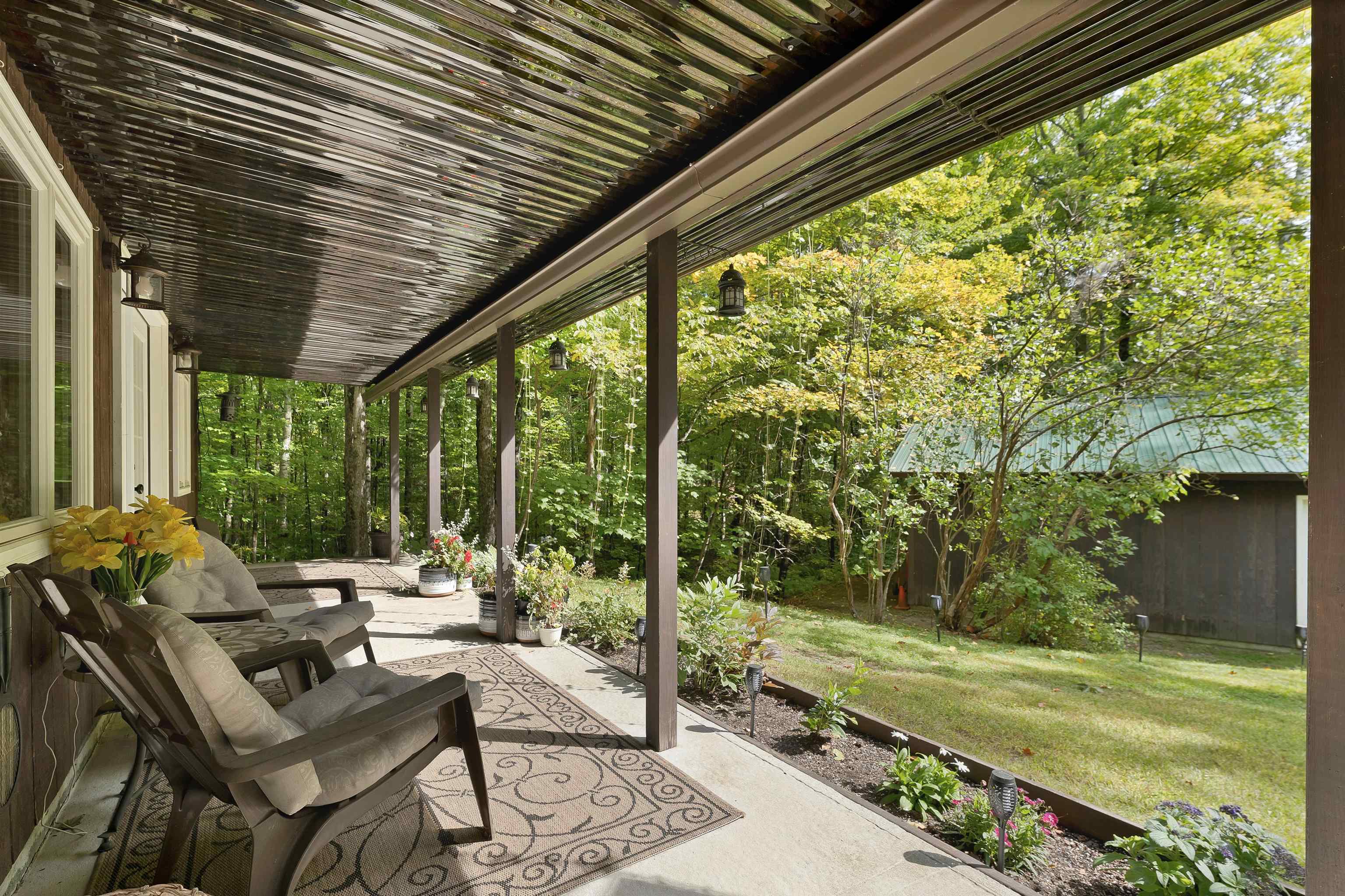 Covered porch with cushioned chairs and potted flowers overlooking a leafy yard and small outbuilding.