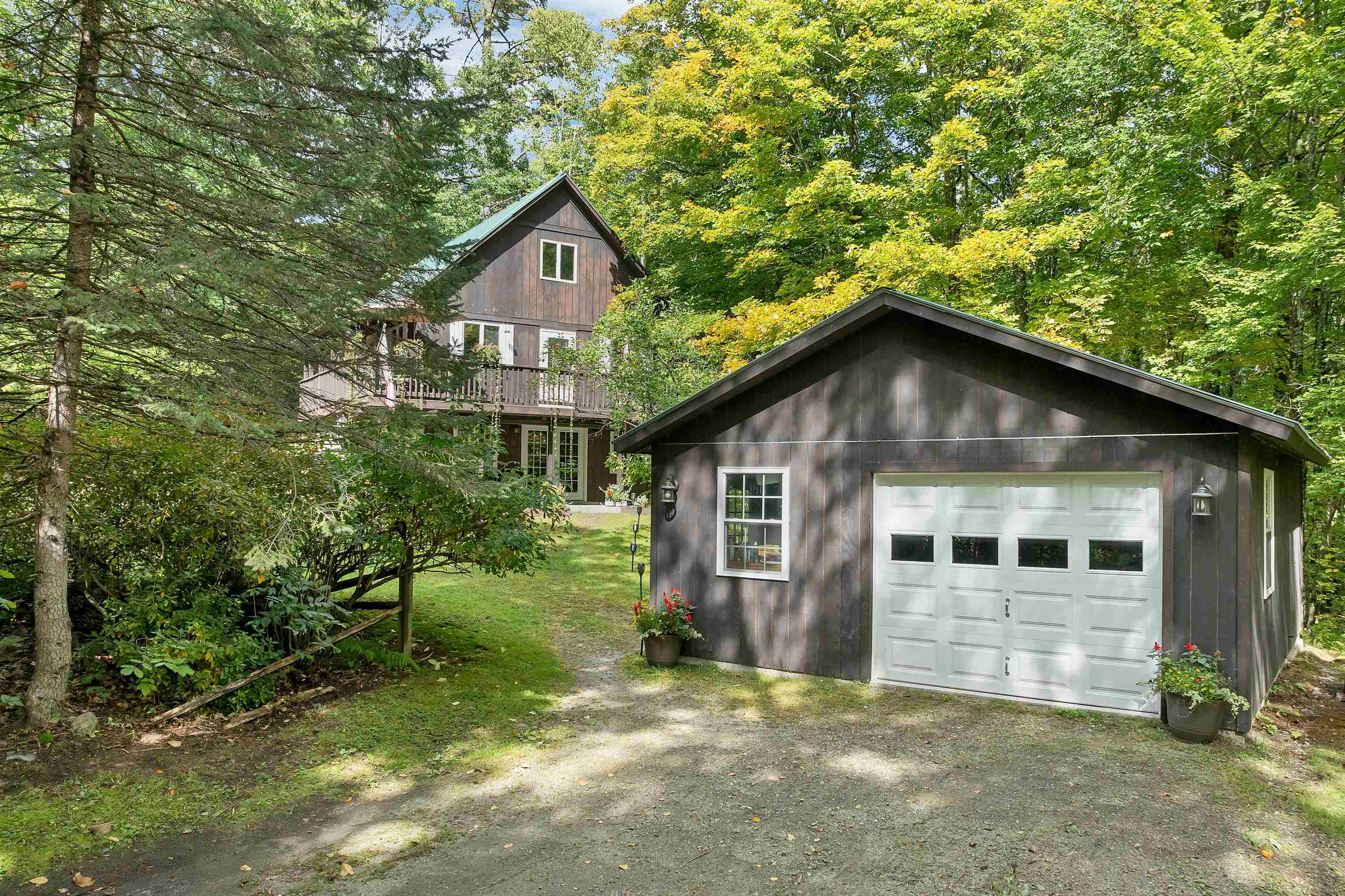 Wood-sided house and detached garage among trees, with a gravel driveway and front deck.