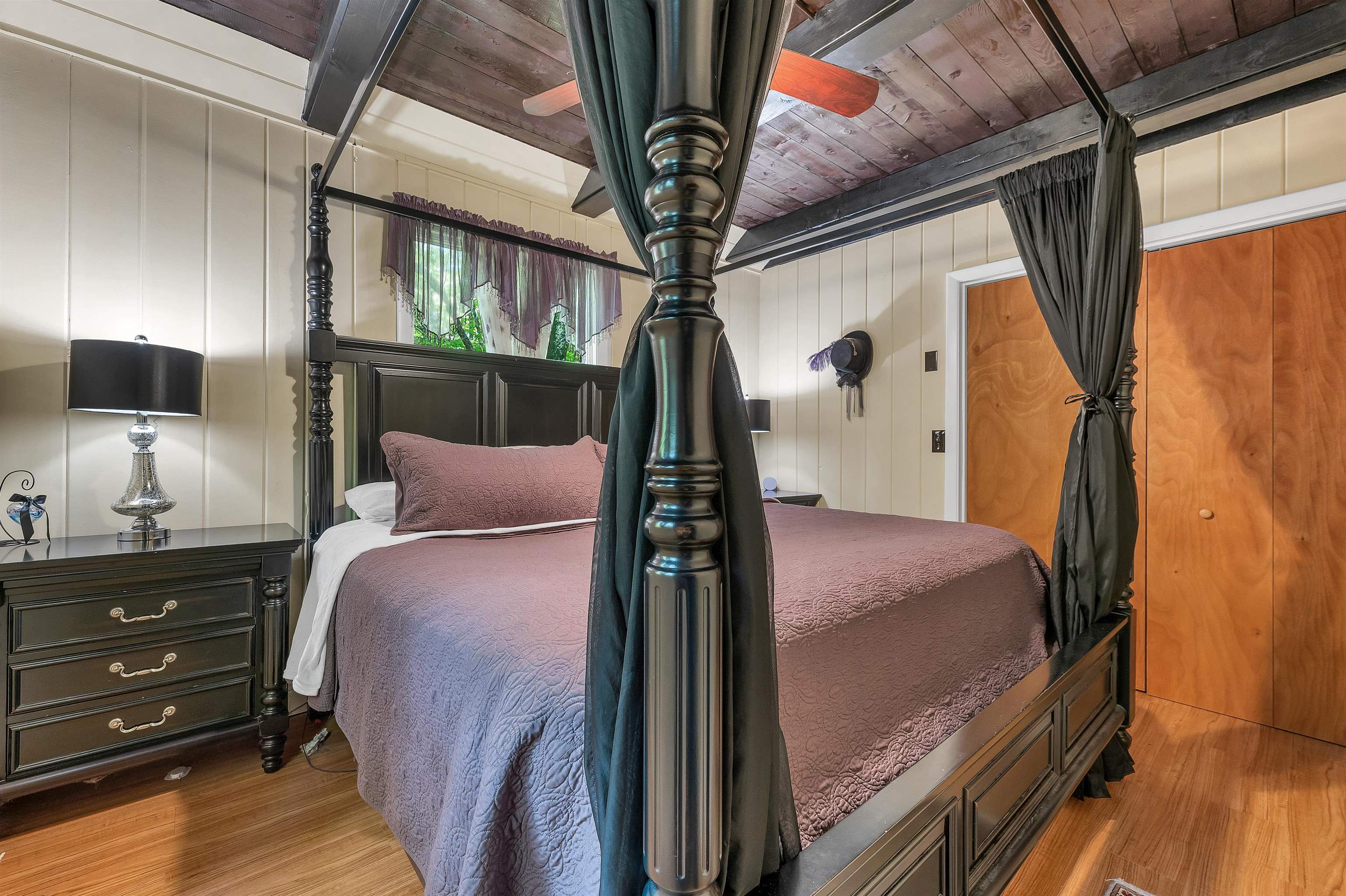 Bedroom with a dark four-poster bed, mauve quilt, exposed wood ceiling, and hardwood floor.