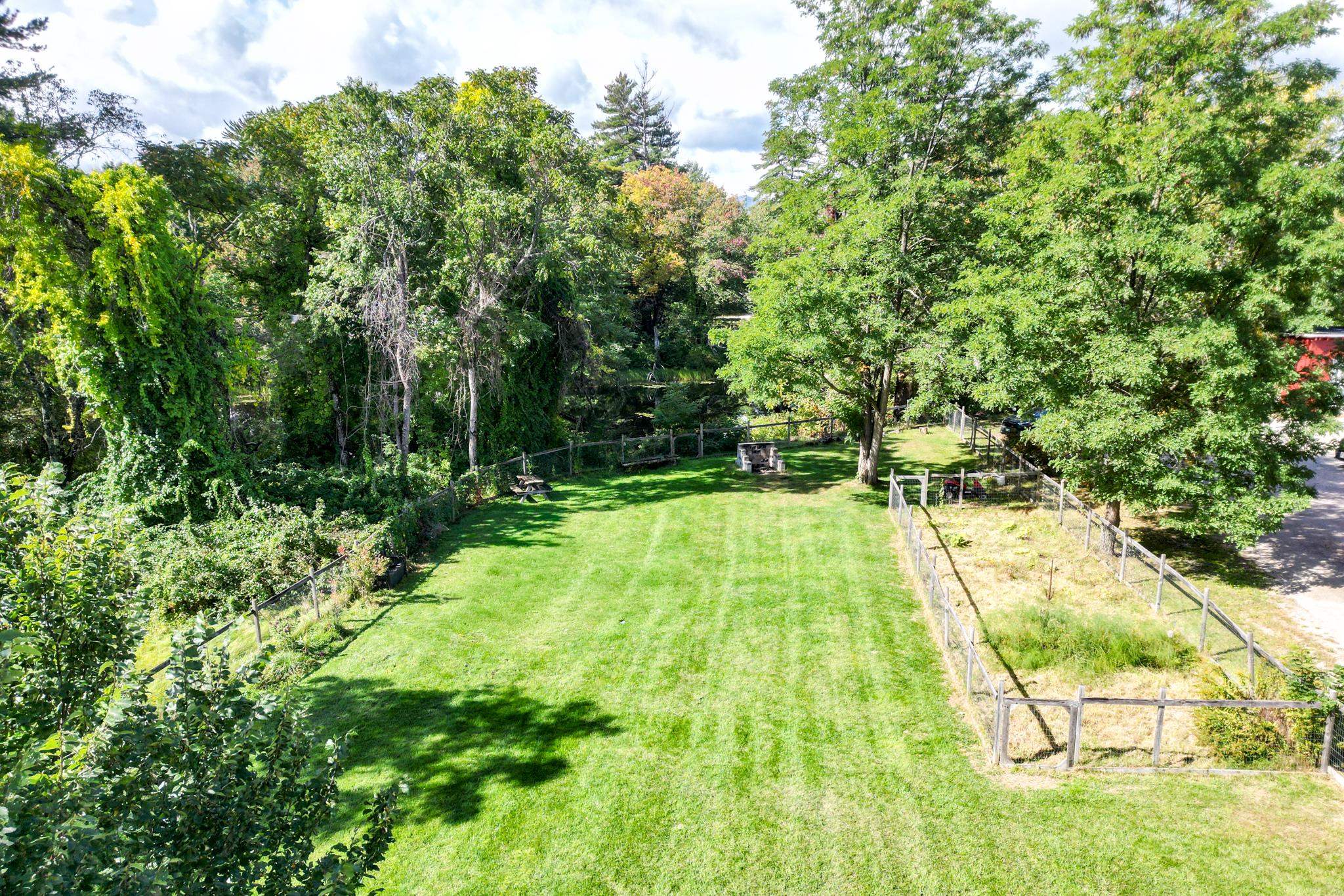 Spacious tree-lined backyard with a broad lawn, fenced garden, picnic table, and stone fire pit.